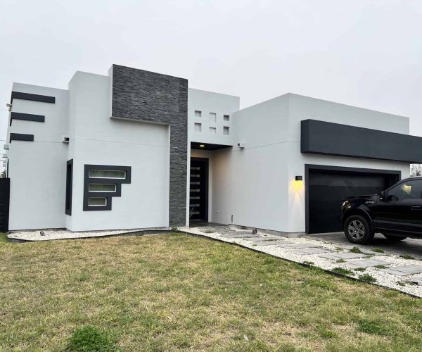 Modern white house with geometric architecture, gray stone accents, and horizontal windows. A black truck is parked on a stone driveway. Overcast sky.