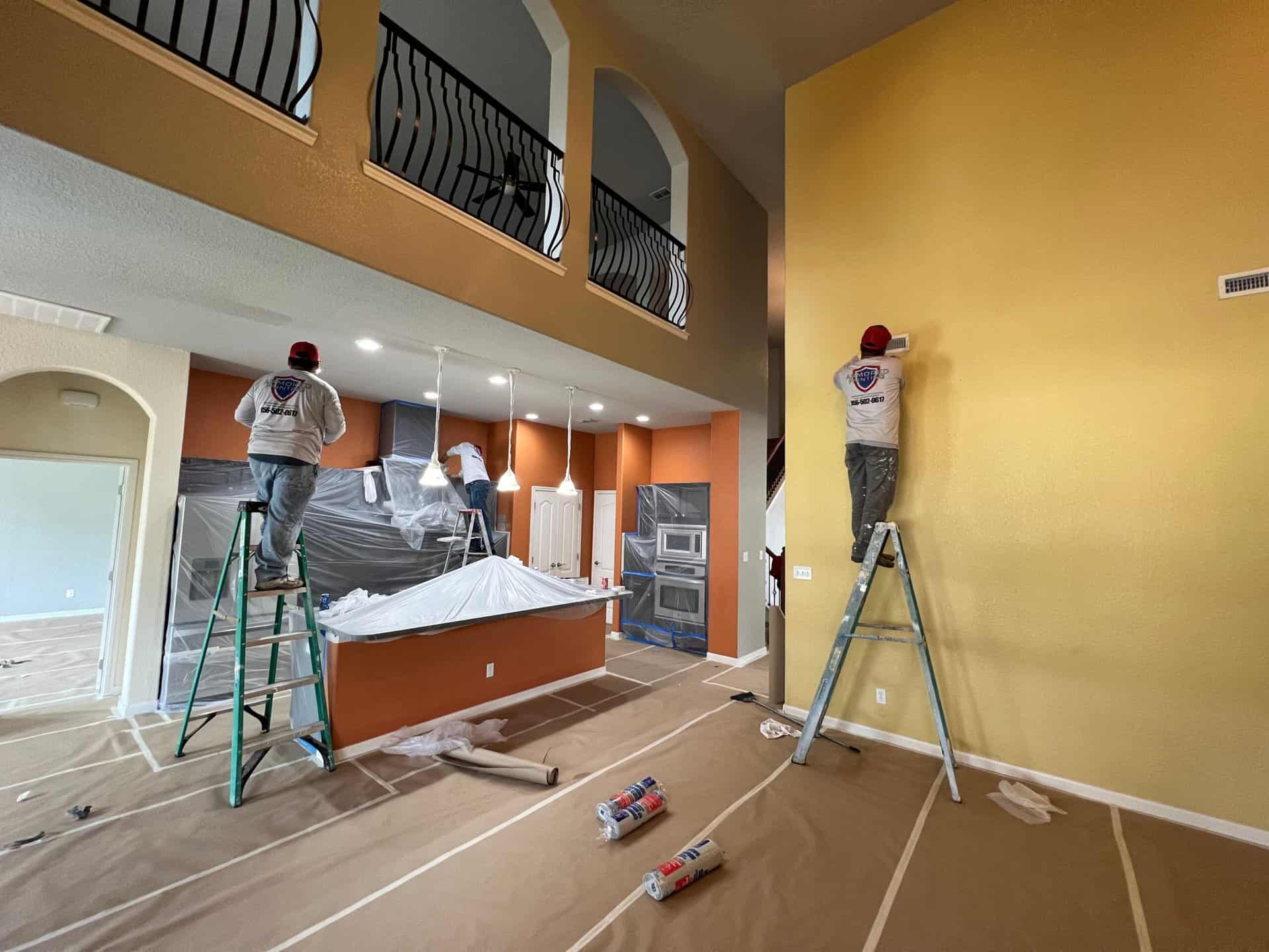Three workers on ladders painting a spacious, high-ceiling room with covered floors and furniture. The walls are yellow and the mood is industrious.
