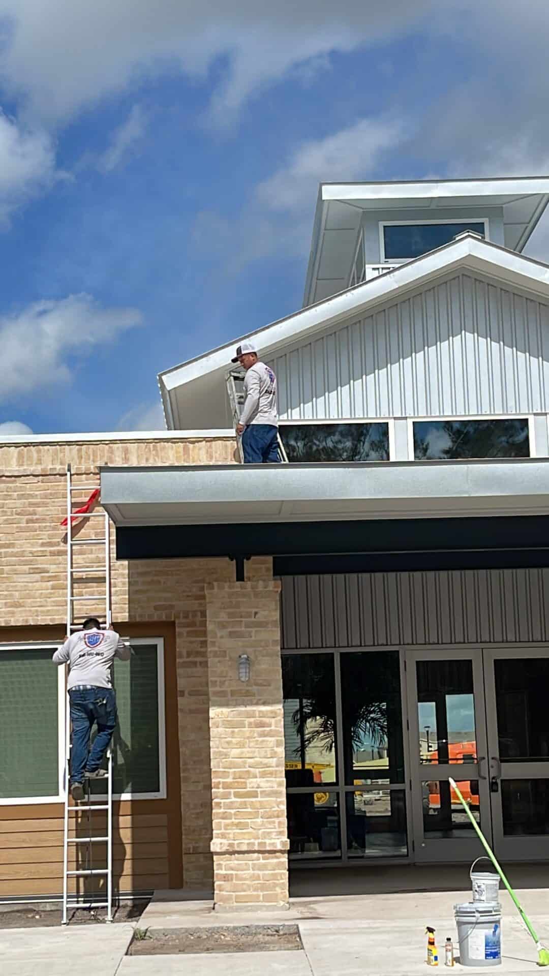 Two workers in matching shirts are on ladders against a building. One is on the roof; the other climbs up. The sky is blue with clouds, giving a busy and industrious tone.