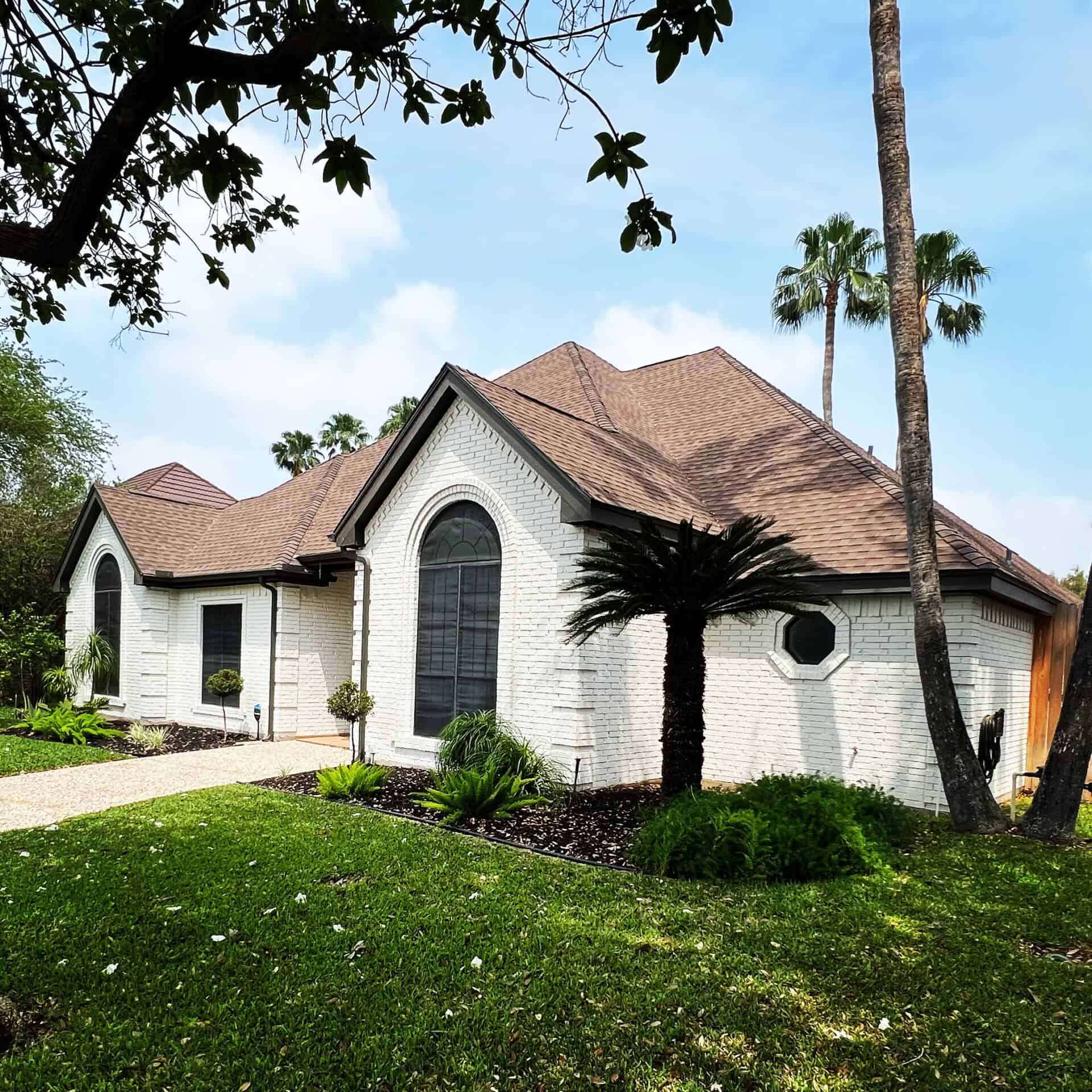 A white brick house with arched windows and a brown roof is surrounded by lush greenery. Tall palm trees stand against a clear blue sky, creating a tranquil vibe.