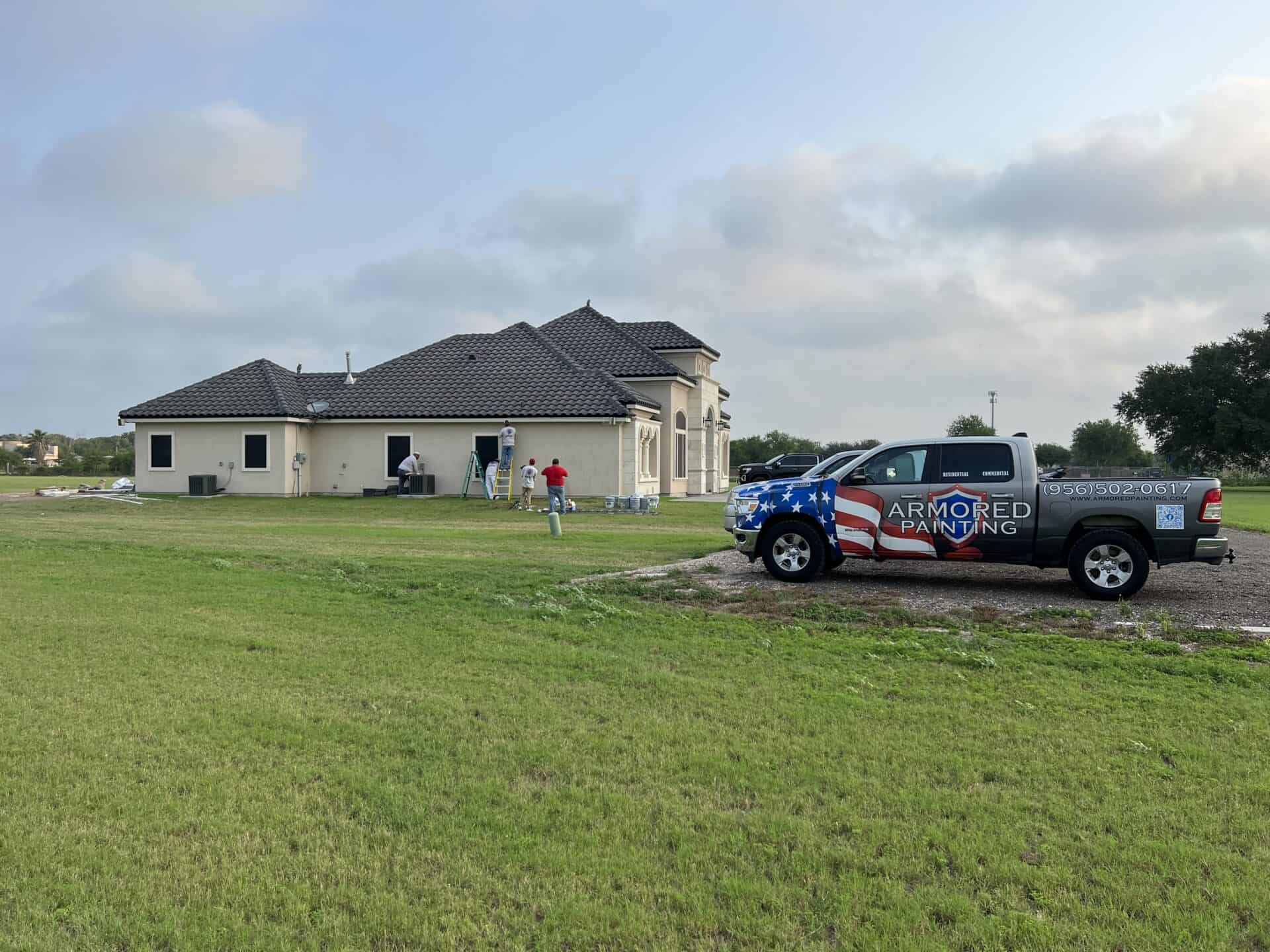 A truck with "Armored Painting" logo, featuring an American flag design, is parked in front of a beige house. Painters in work attire are on ladders painting the home's exterior. The scene conveys a sense of productivity on a grassy lawn under a partly cloudy sky.