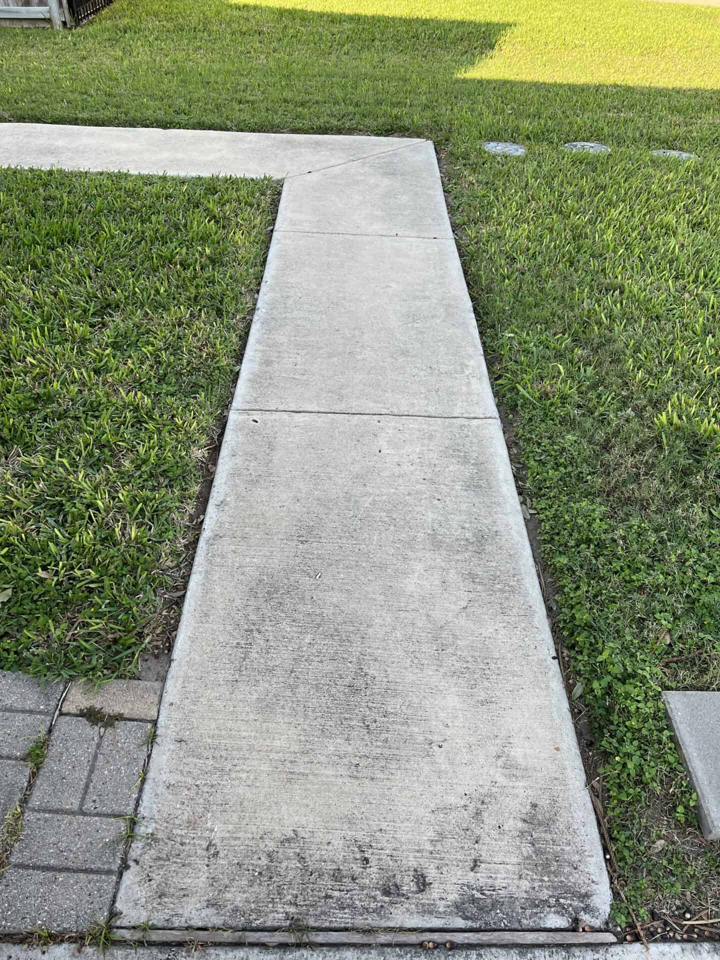 Concrete pathway curving through green grass, bordered by gray bricks at the bottom. The scene is bright, evoking a peaceful suburban setting.