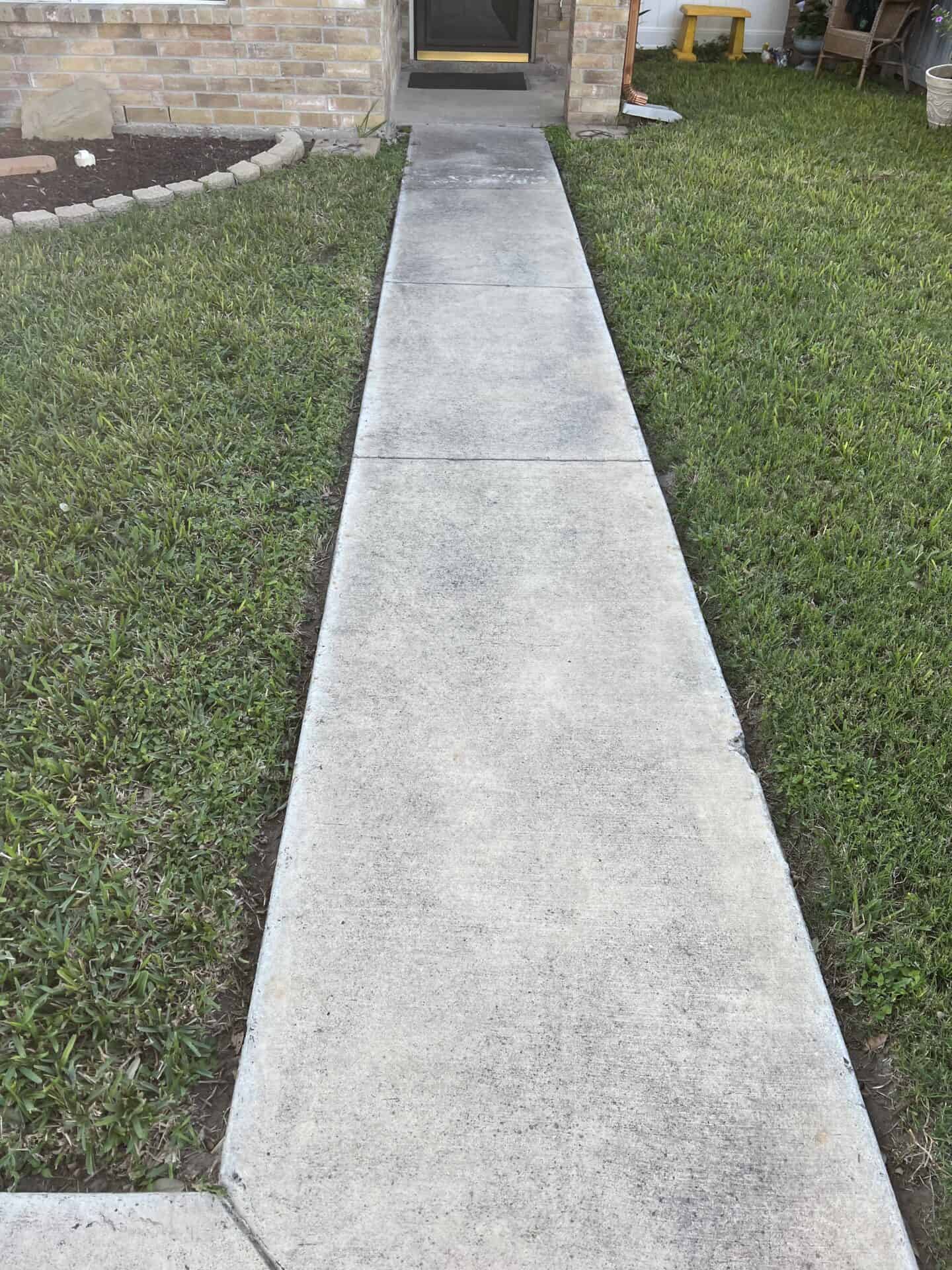 Concrete pathway curving through green grass, bordered by gray bricks at the bottom. The scene is bright, evoking a peaceful suburban setting.