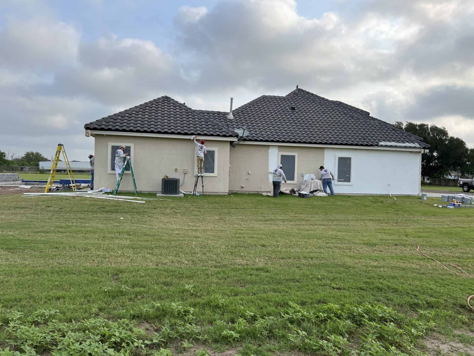 Several workers, wearing white clothes and using ladders, are painting the beige exterior of a house with dark roof tiles. The mood is industrious.