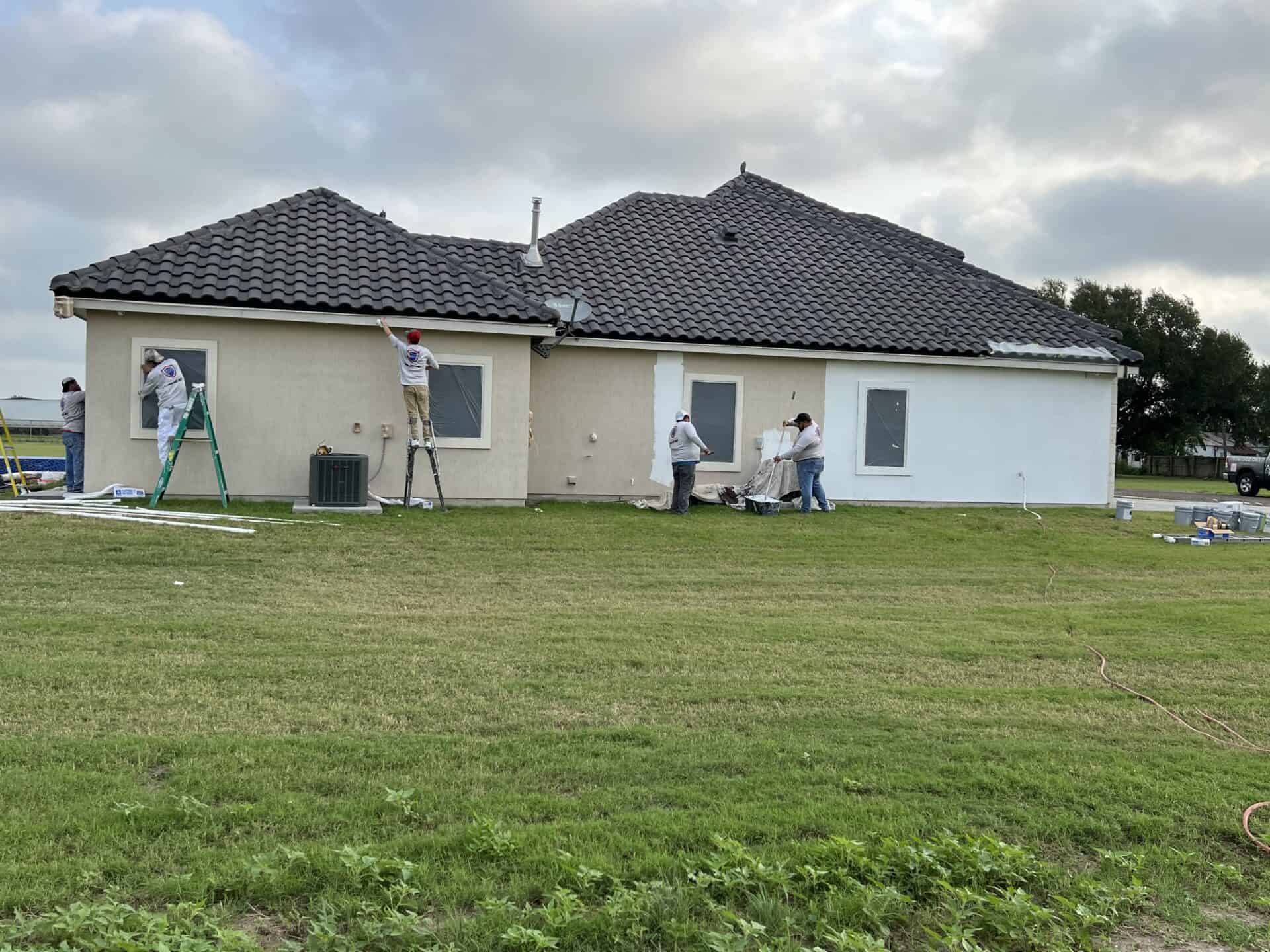 Workers painting the exterior of a beige house with dark shingles. Ladders and painting supplies are visible on the lawn under a cloudy sky.