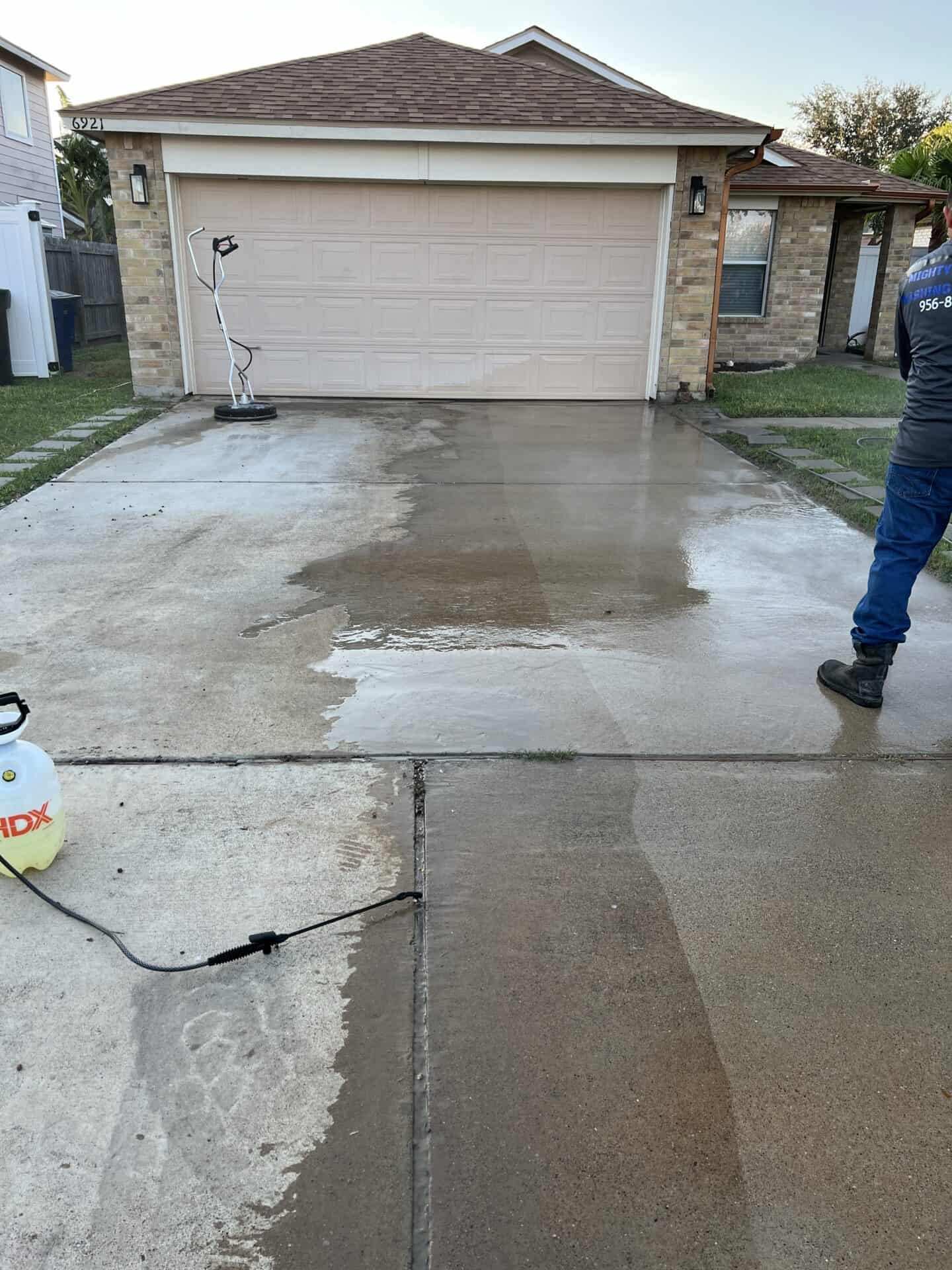 A driveway being pressure washed, showing a stark contrast between the cleaned and dirty sections. A worker is visible with equipment nearby.
