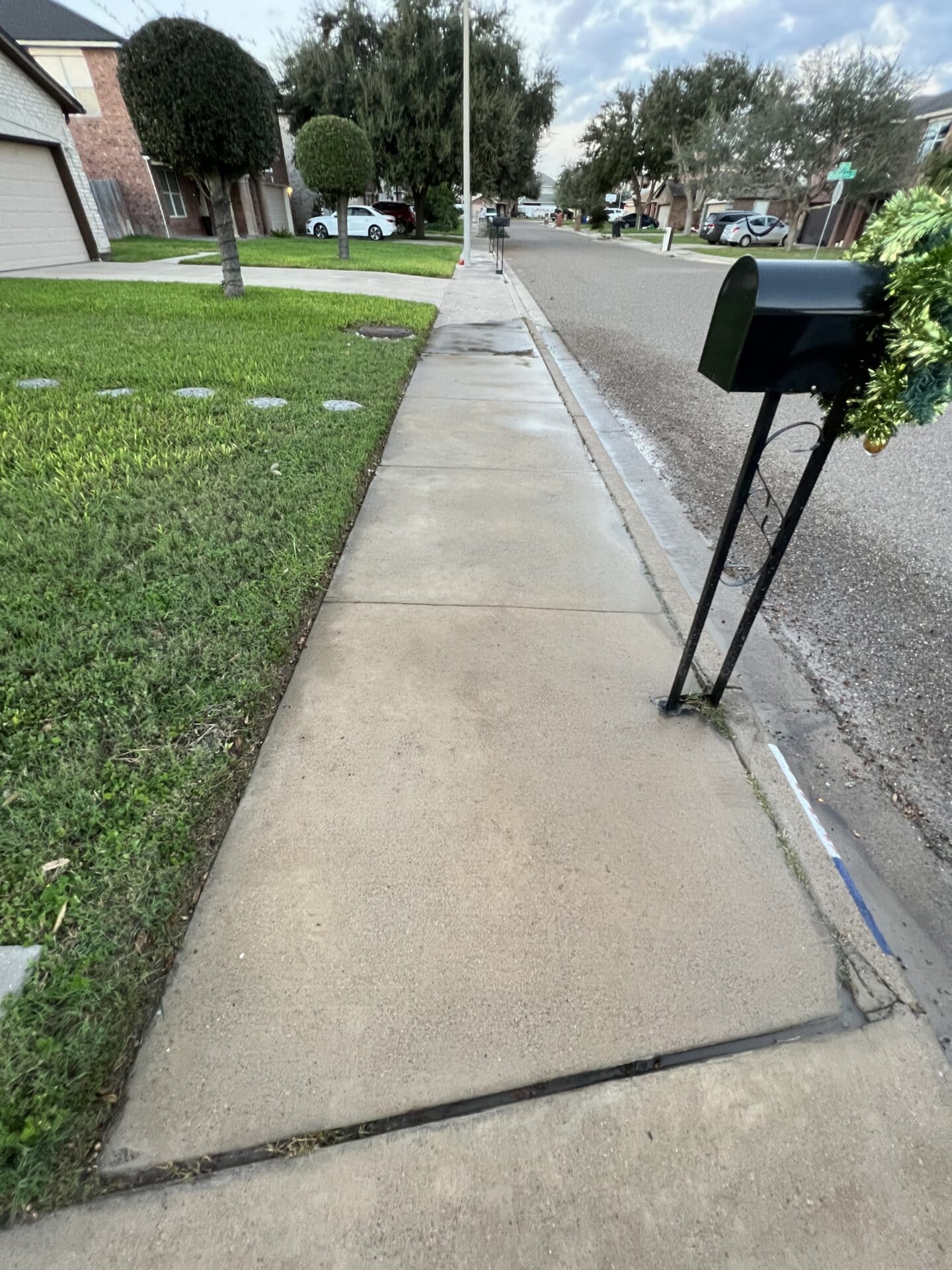 Sidewalk view in a suburban neighborhood with green lawns, trimmed trees, and houses. A festive mailbox with garland is in the foreground. Cloudy sky above.