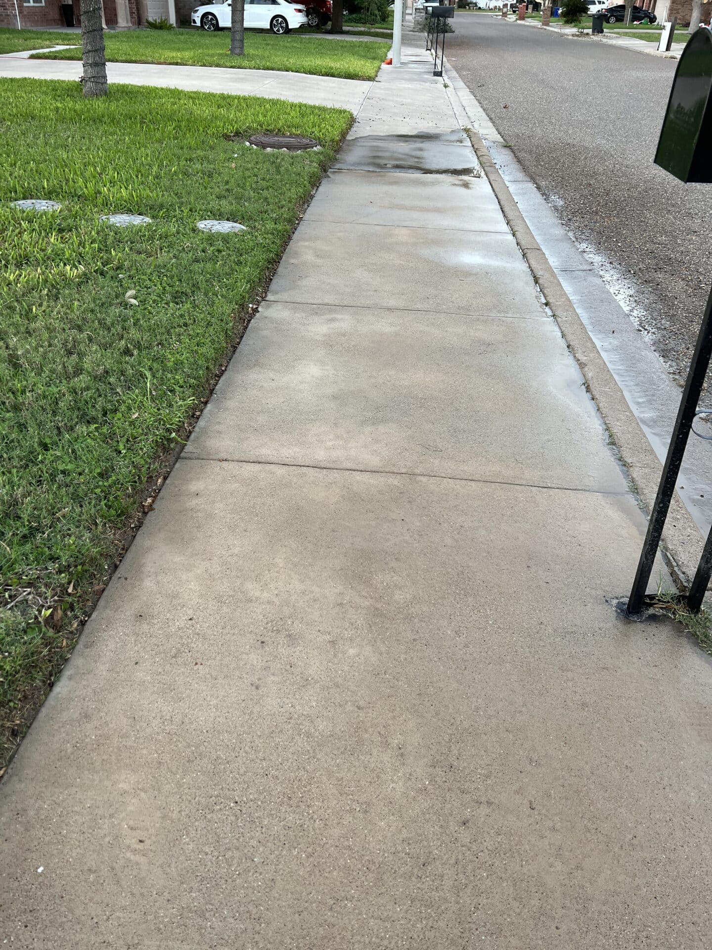 A straight, wet sidewalk beside a grassy lawn leads up to a residential street. A white car is parked nearby, reflecting a calm suburban scene.