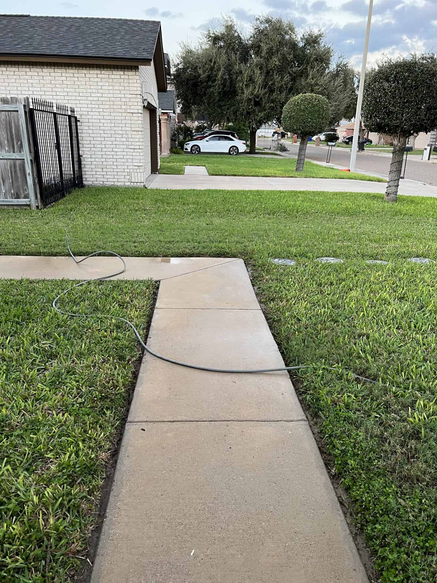 A concrete path runs through a neat green lawn with a coiled hose laying across it. Trees and a parked car in the background under a lightly cloudy sky.