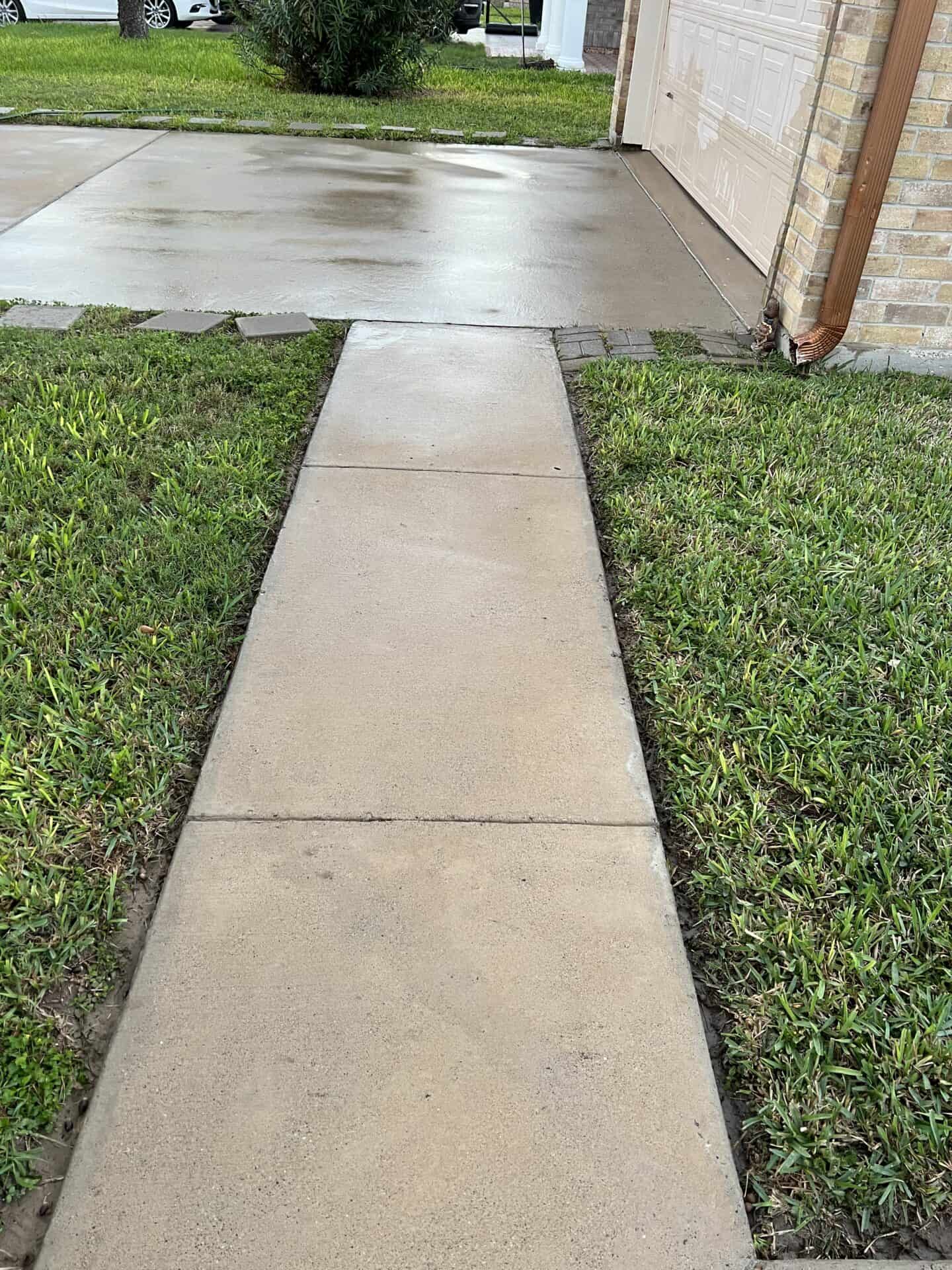 A clean concrete walkway leads to a wet driveway beside a brick house wall. Grass lines both sides, and a white car is visible in the background.