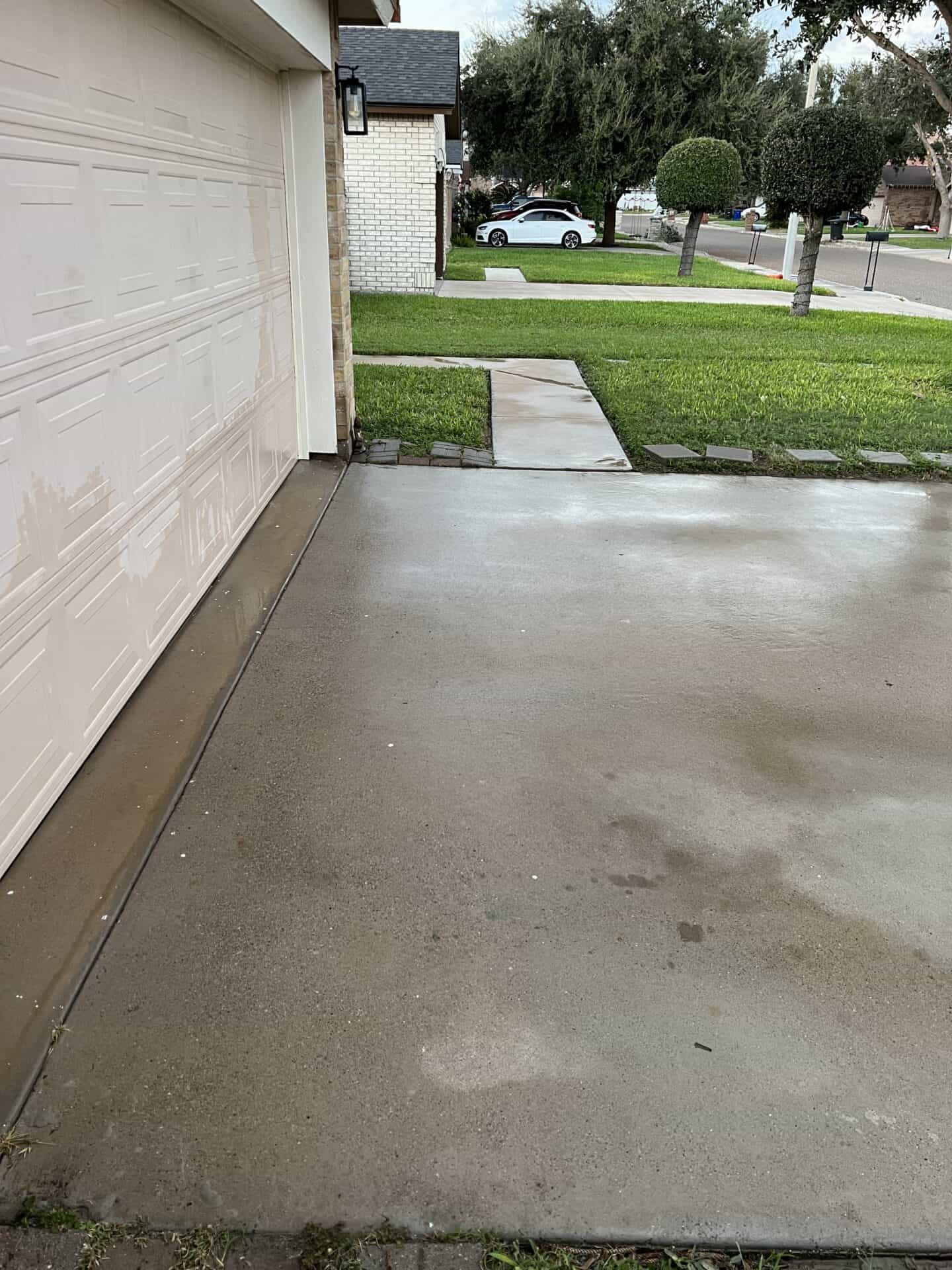 A wet concrete driveway leads to a closed garage door. A grassy lawn and a small tree are on the right, with a white car parked on the street. Overcast sky adds a calm mood.