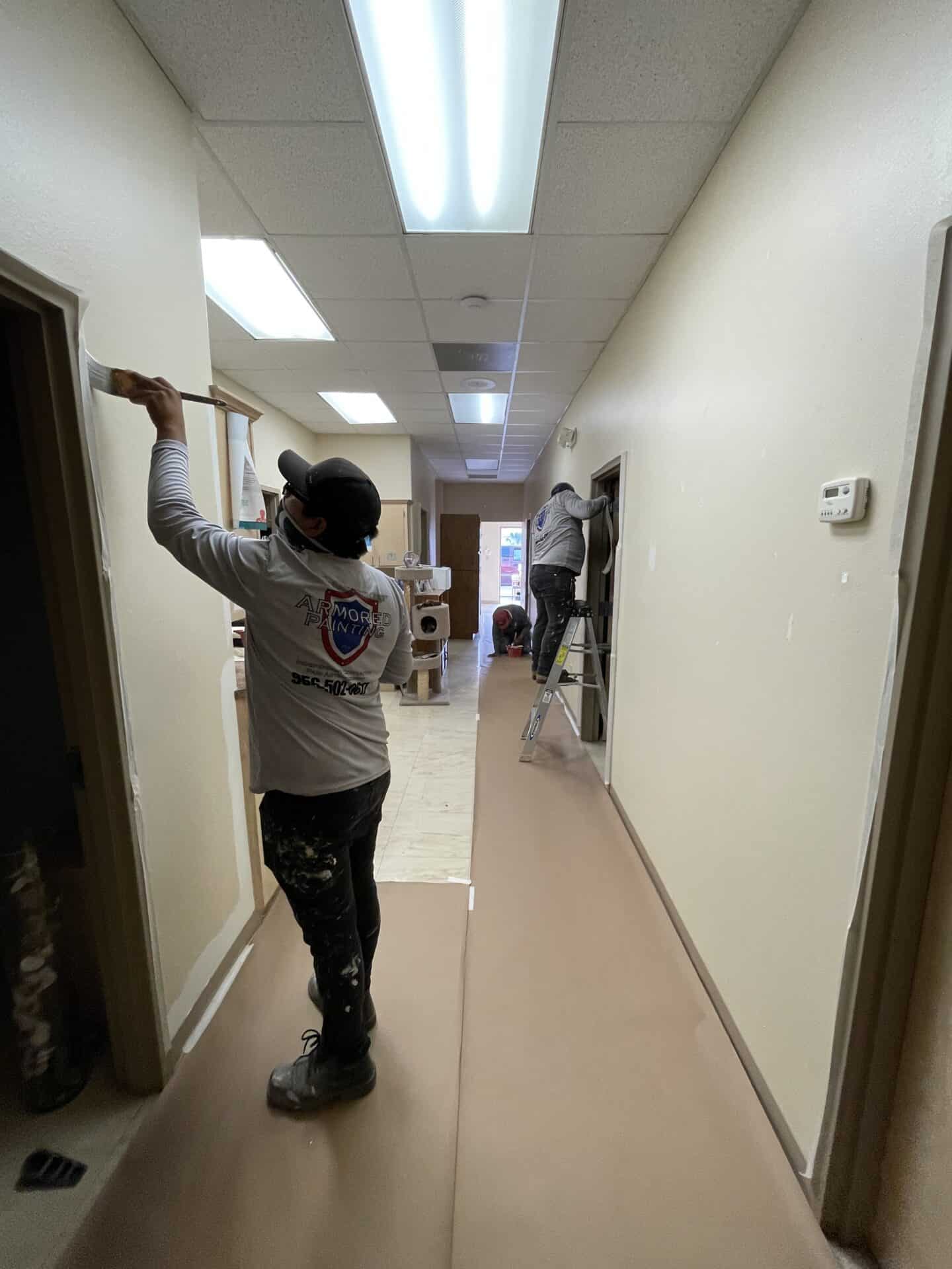 Hallway painting in progress. Three workers in uniforms, one on a ladder, apply fresh paint. Bright overhead lights and a calm atmosphere dominate.