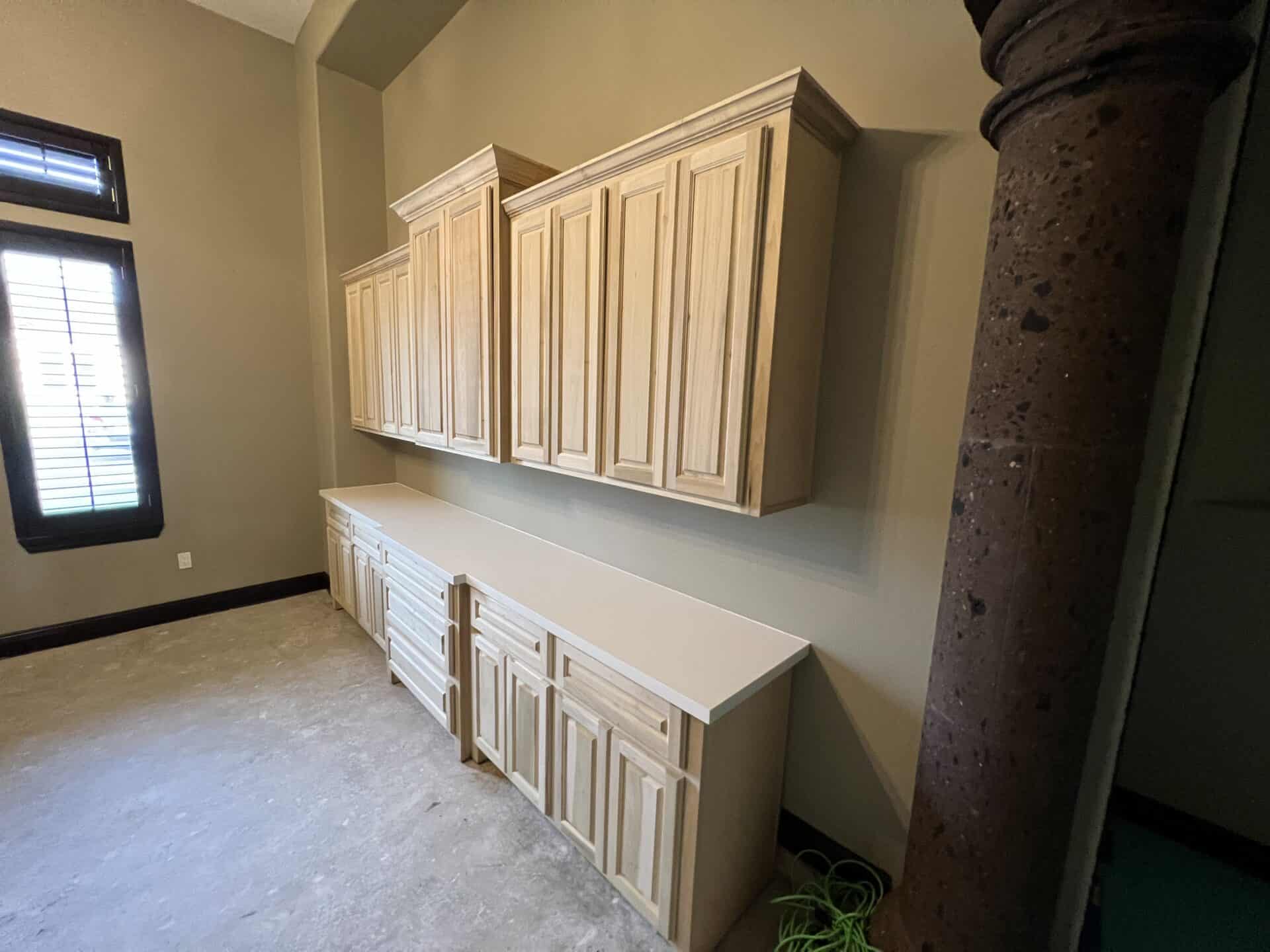 Room with light wood cabinets against a beige wall, featuring both overhead and base storage. A column and window with shutters are visible, creating a calm ambiance.