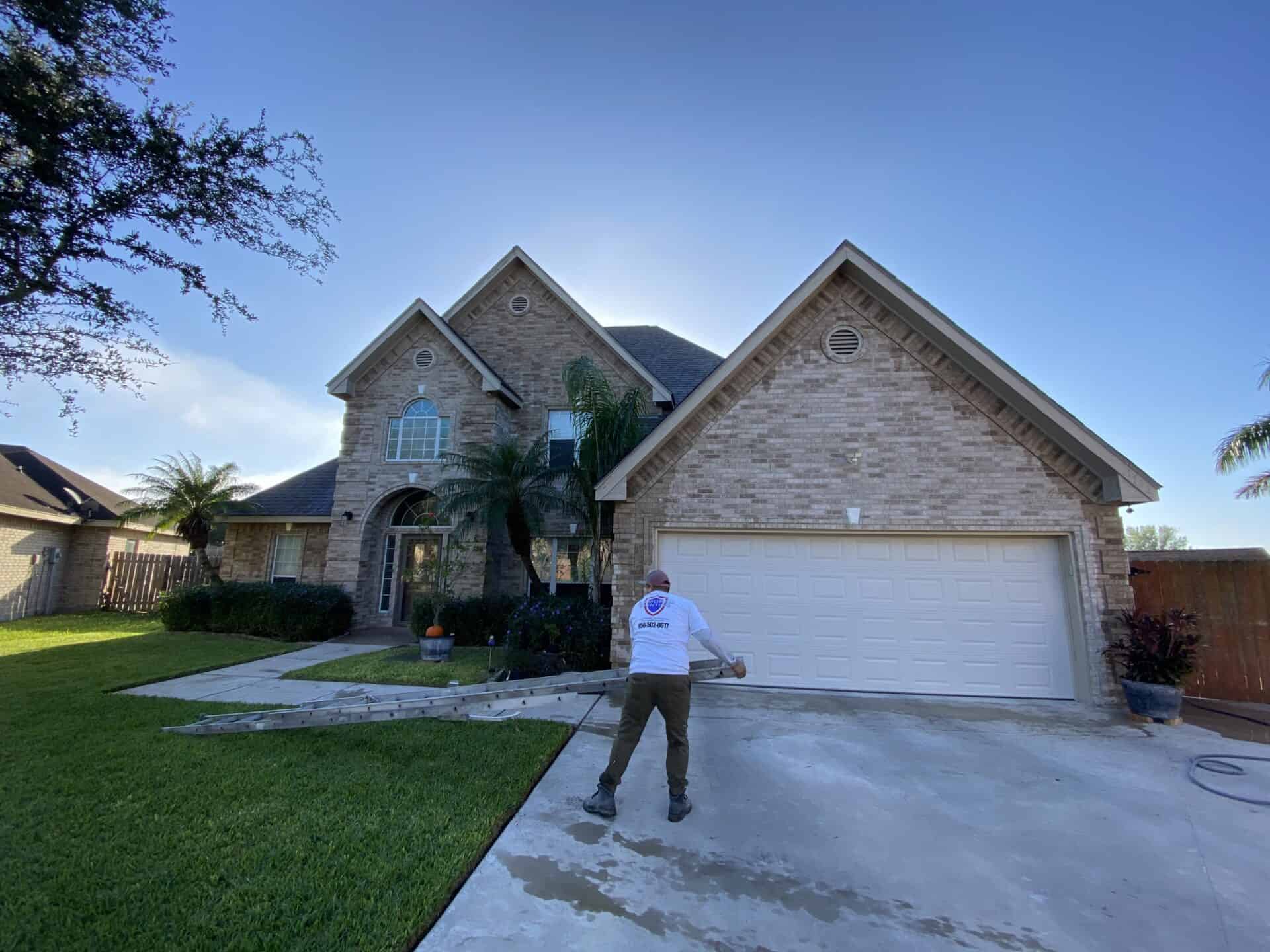 Two-story brick house with a large garage and manicured lawn. A person cleans the driveway with power-washing equipment under a clear blue sky.
