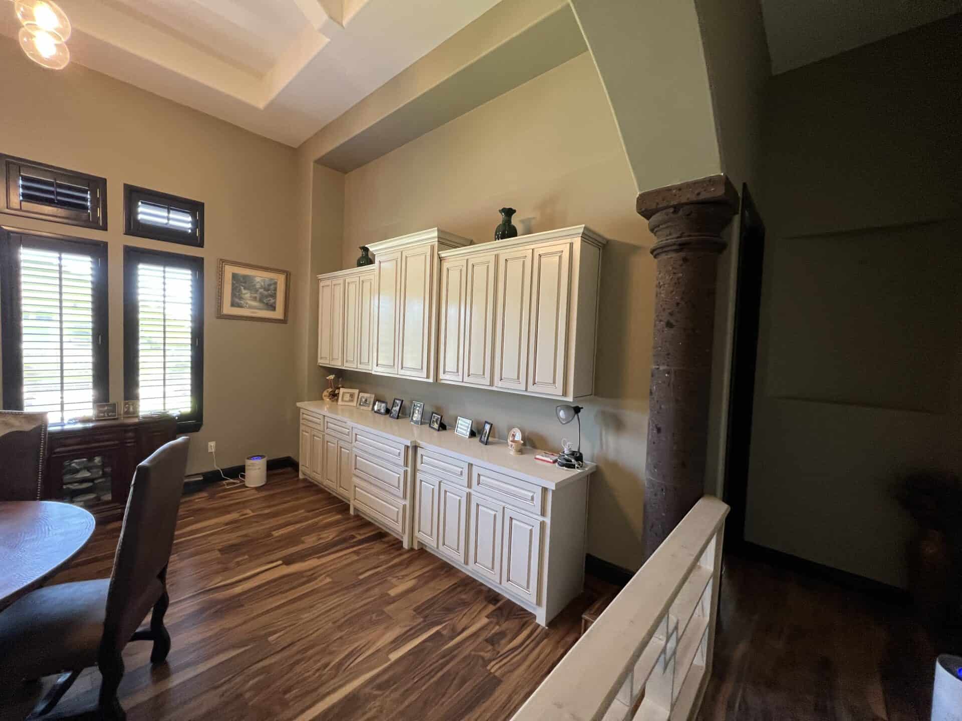 A cozy room with wooden floors, featuring a round dining table, chairs, and a wall of white cabinets displaying framed photos. Soft natural light filters through tall windows, creating a warm, inviting atmosphere.