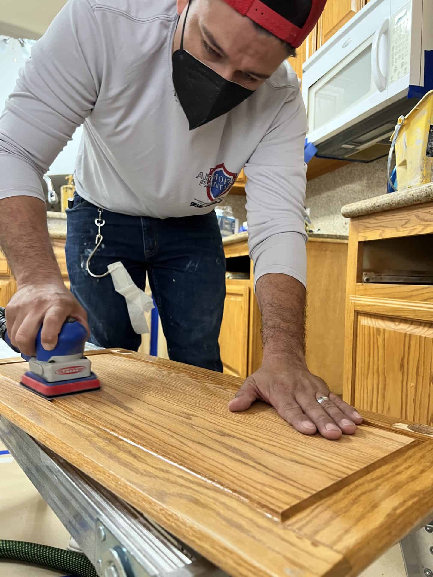 A person wearing a red cap and black mask sands a wooden cabinet door in a kitchen. They focus intently, creating a sense of concentration.
