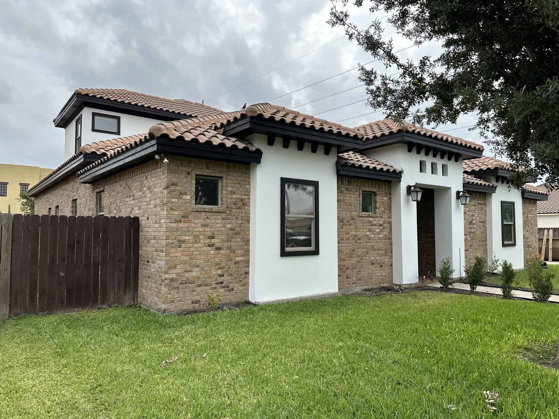 Single-story house with beige brick and white stucco exterior, featuring a red-tiled roof. The lush green lawn and overcast sky create a calm atmosphere.