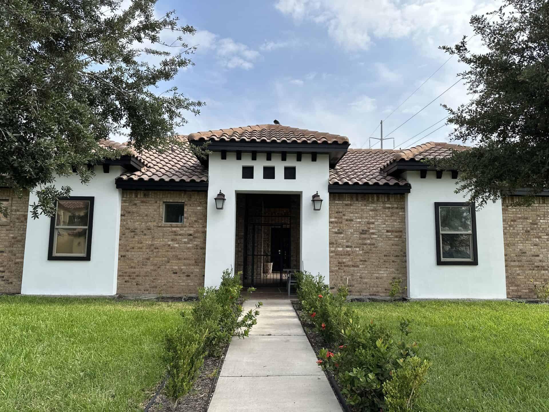 Single-story house with a mix of white stucco and brick walls, brown tiled roof, and a central entrance. Framed by green grass and trees under a clear sky.