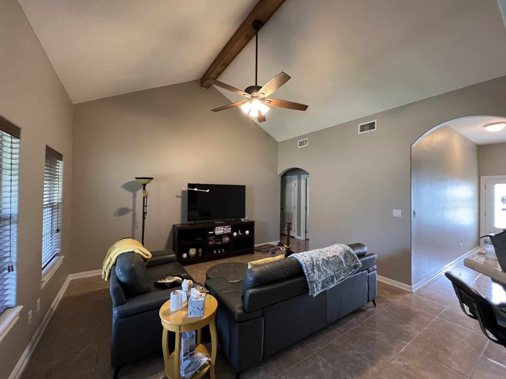 Cozy living room with high ceiling and exposed beam. Features a black leather sofa, TV, and bright ceiling fan. Warm lighting and neutral tones create a calm atmosphere.