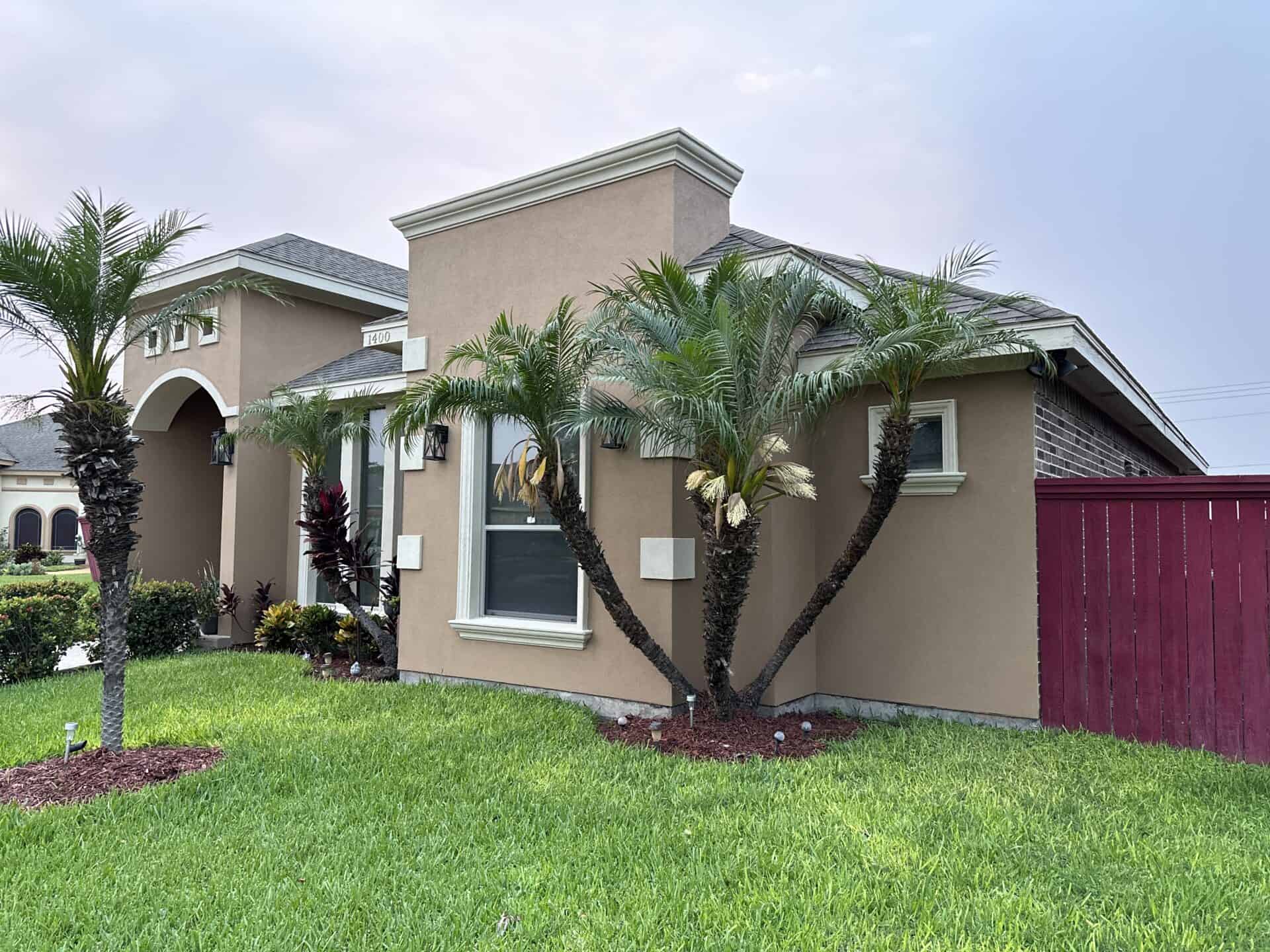 Single-story house with beige stucco, palm trees, arched entryway, and red wooden fence. Vibrant green lawn and landscaped garden enhance charm.