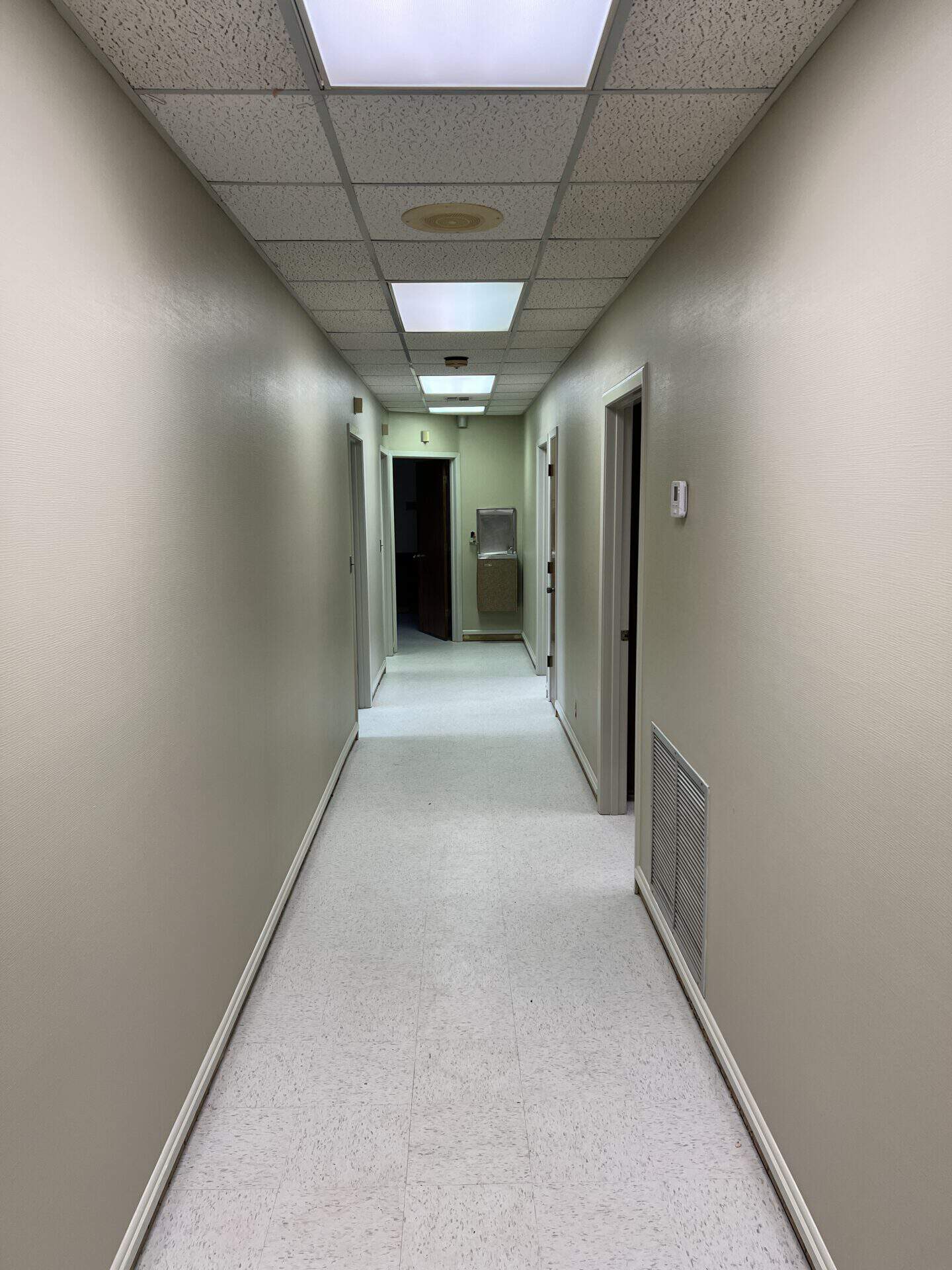 A narrow hallway with beige walls and ceiling tiles is brightly lit by overhead fluorescent lights. Doors line each side, leading to unseen rooms. The atmosphere feels sterile and quiet.