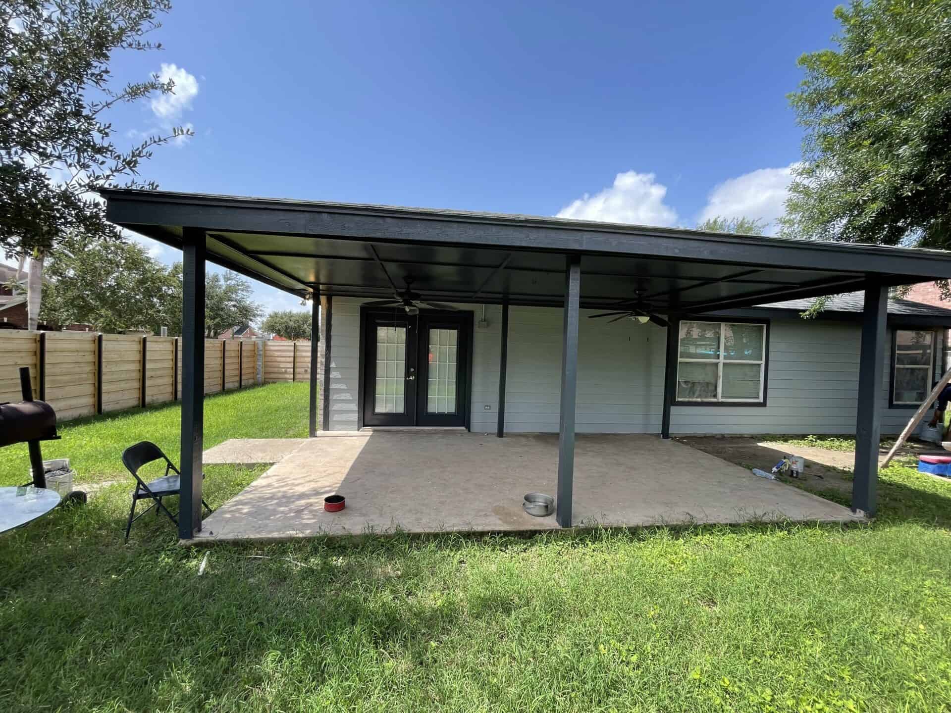 Covered patio with dark wooden beams attached to a gray house. It overlooks a green lawn with a wooden fence. A grill and folding chair are visible.