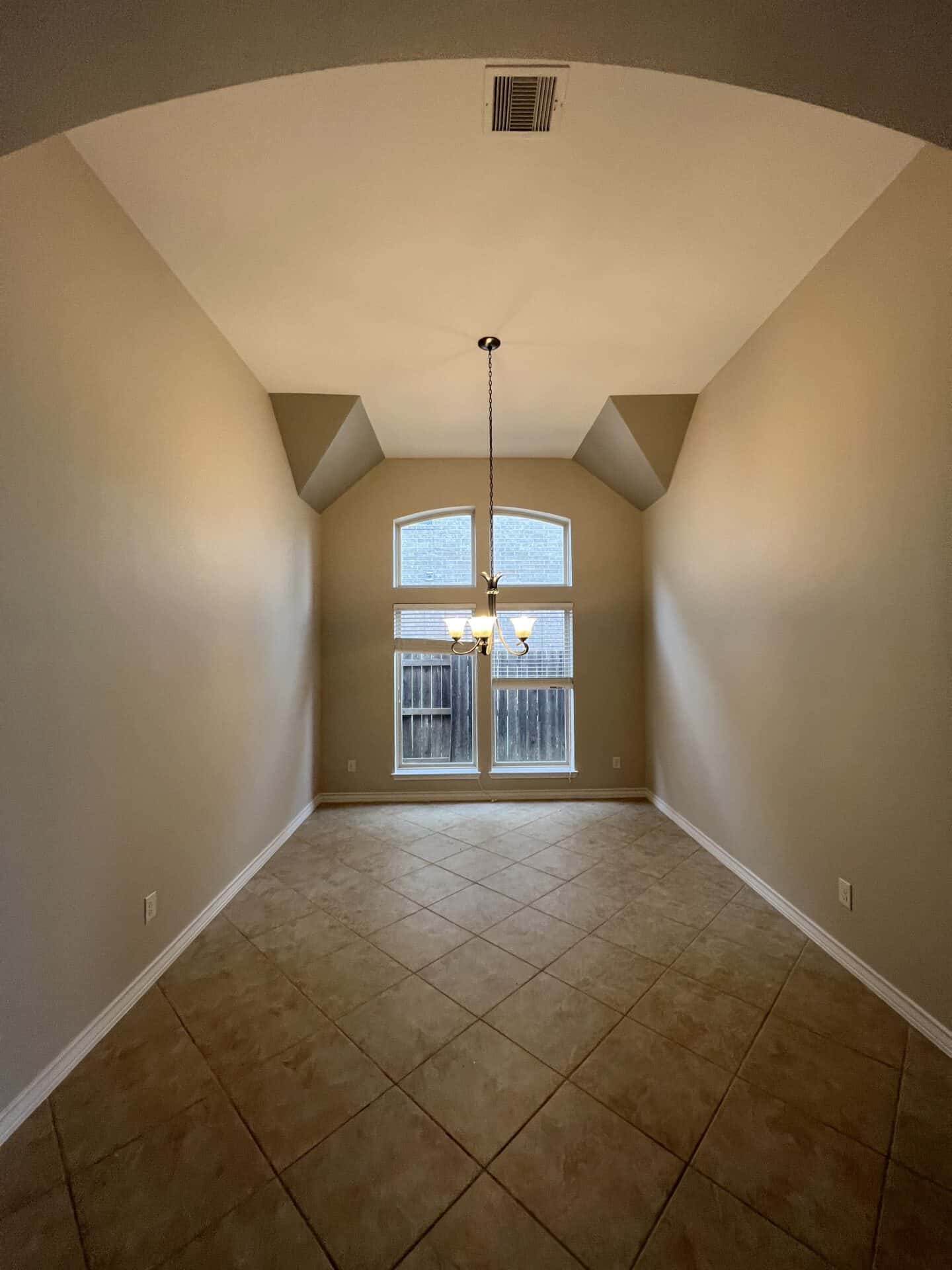 Bright, empty room with beige walls and tiled floor, featuring a high ceiling and arched window. A chandelier hangs from the ceiling, adding a touch of elegance.