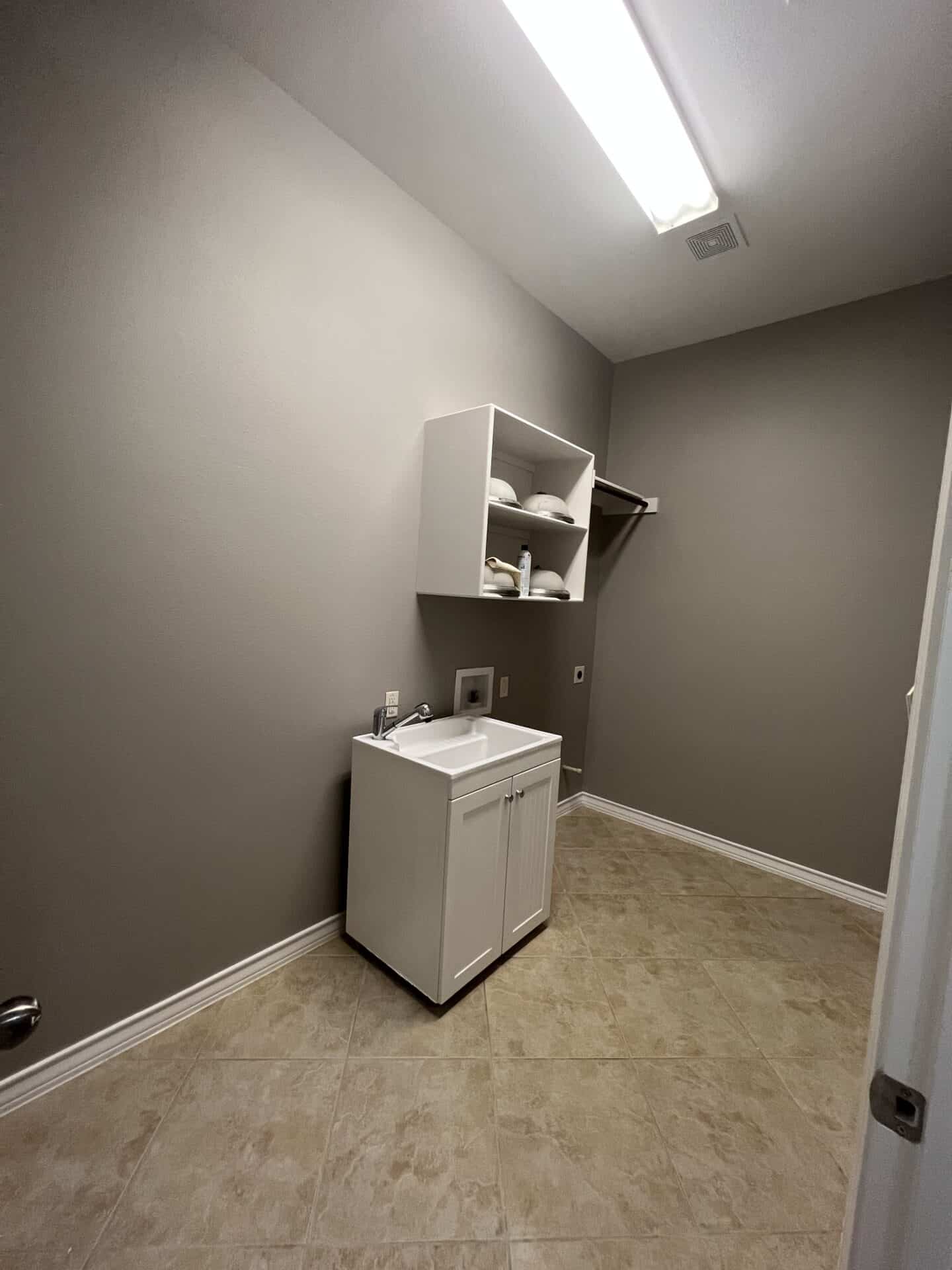 A minimalist laundry room with light beige tiled floor and gray walls. A white sink cabinet is below a wall shelf holding folded towels. Ceiling light illuminates the space.