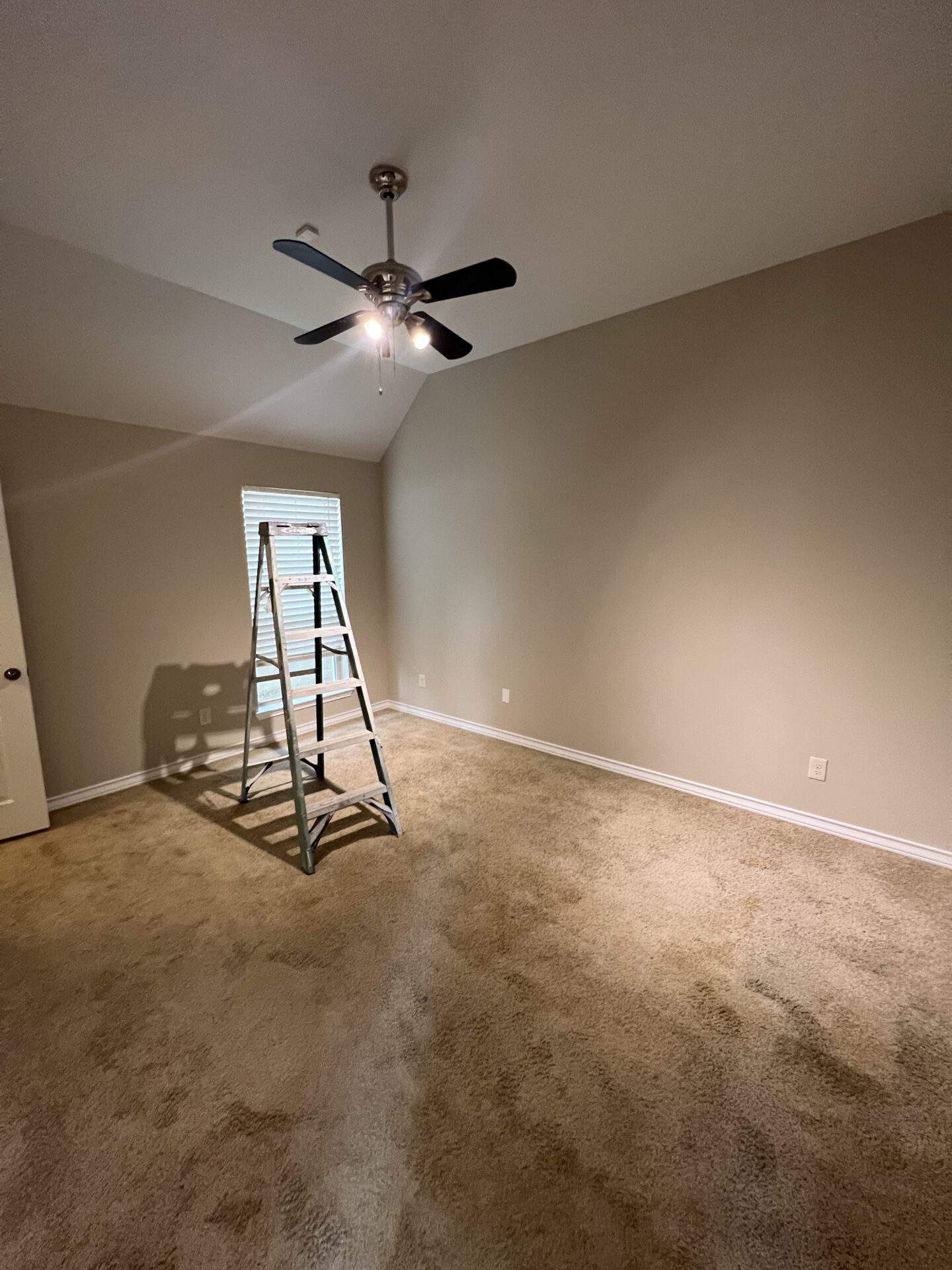 Empty beige room featuring a silver ceiling fan with lights, a ladder under a window with blinds, and soft carpet flooring, conveying a neutral tone.