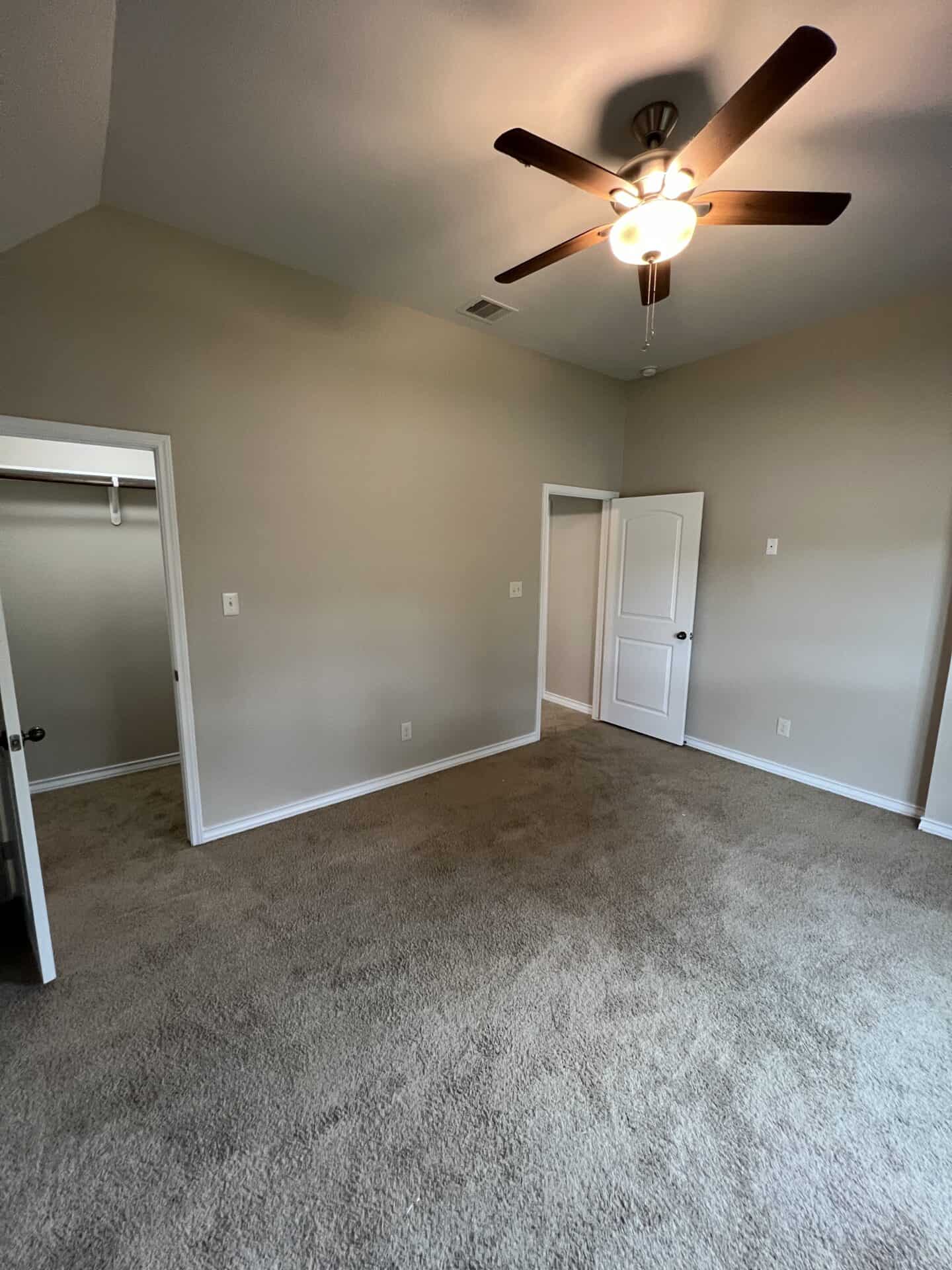 Empty room with beige carpet, light gray walls, and a ceiling fan with lights. Two open doors reveal a closet and another room, creating an inviting and clean atmosphere.