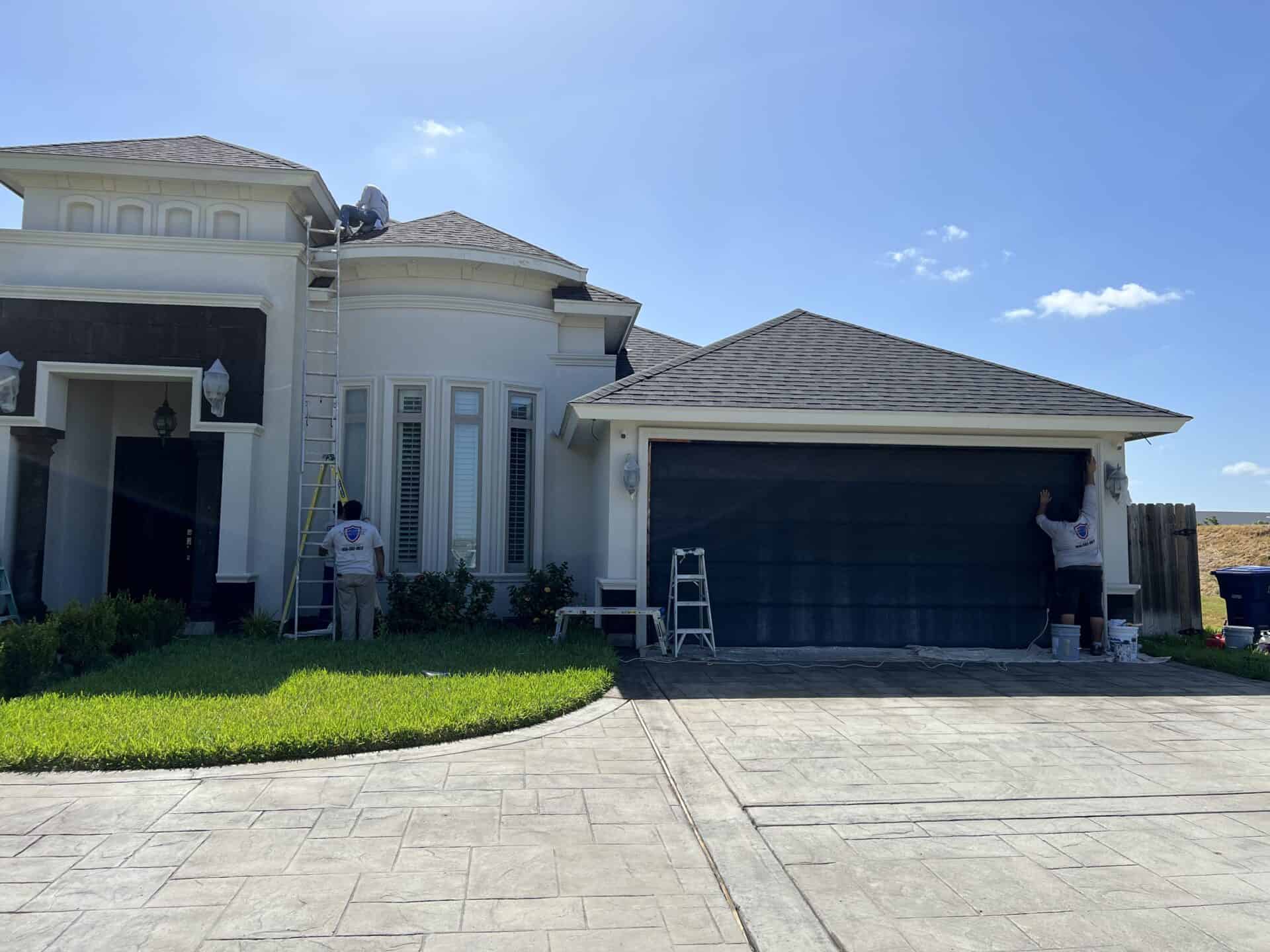A team of workers paints a modern house under a clear sky. One is on a ladder near the roof, while another paints the garage door. The atmosphere is industrious.