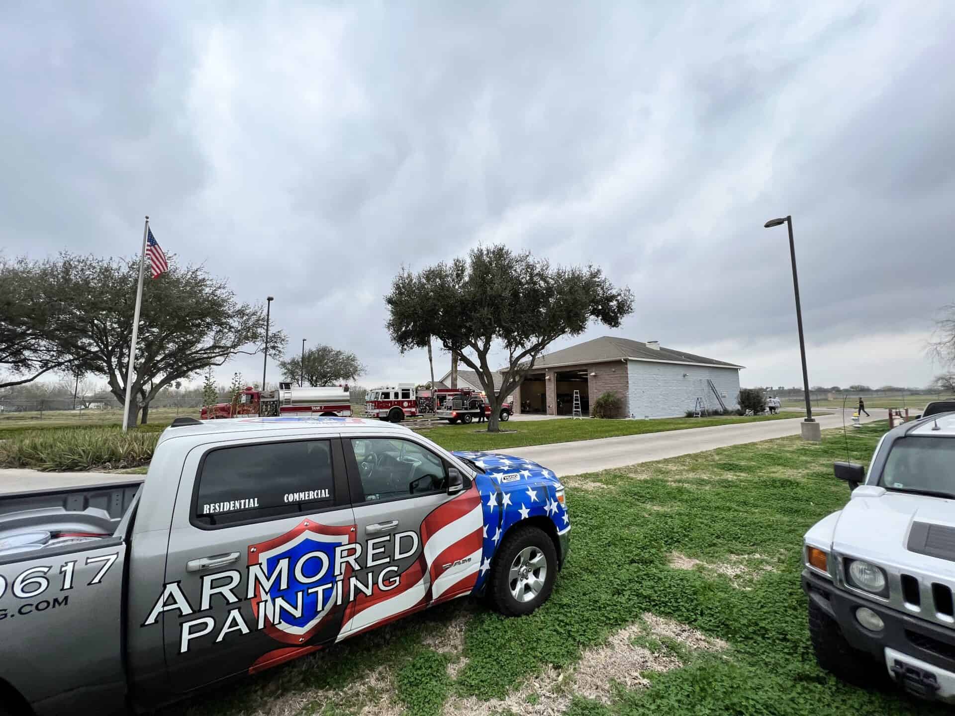 Pickup truck with "Armored Painting" logo parked near a building. Overcast sky with American flag and firefighting vehicles in the background. Calm scene.