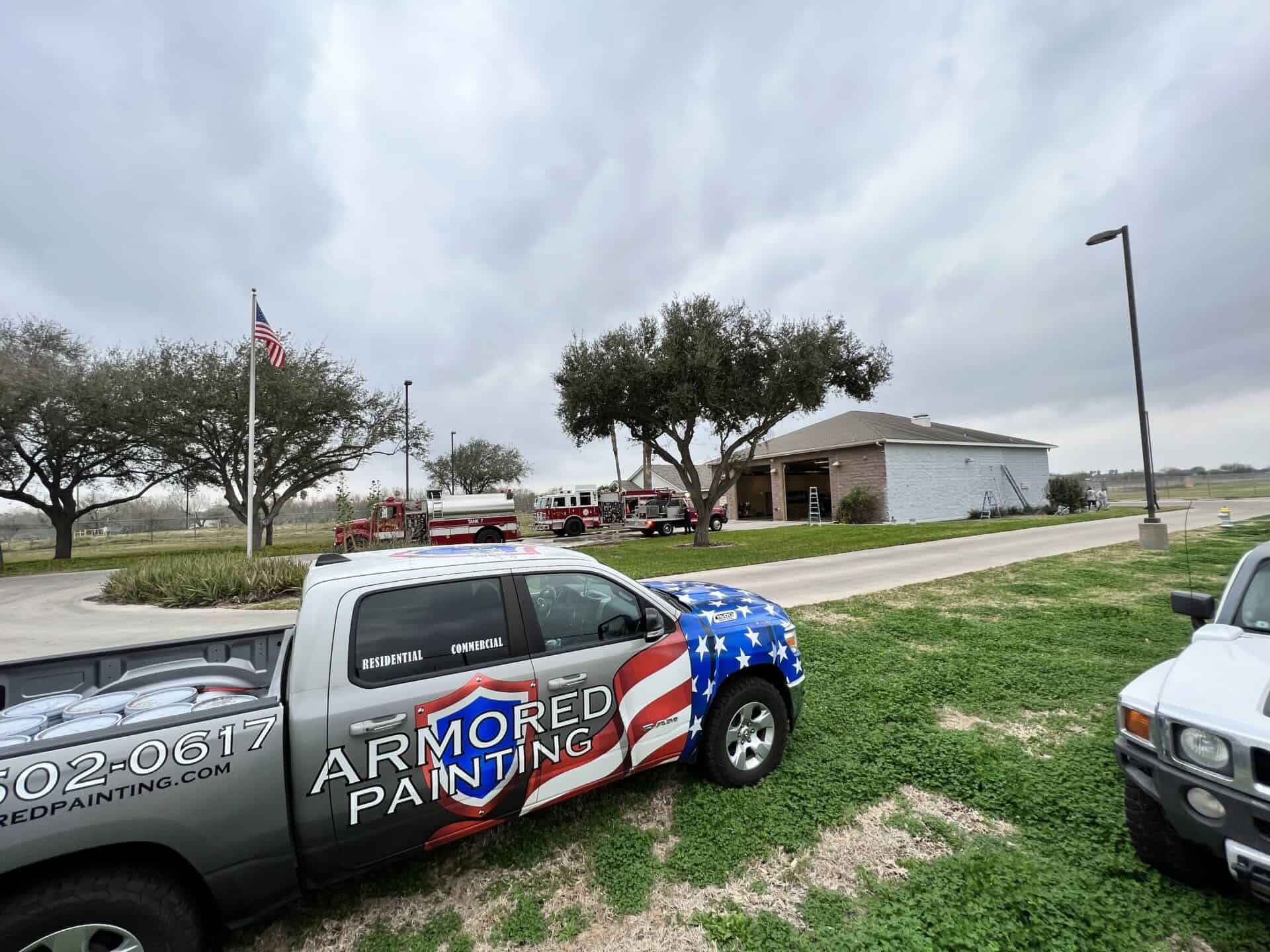 A truck with an "Armored Painting" logo and stars and stripes design is parked on grass. Nearby, fire trucks are outside a fire station. The sky is overcast.