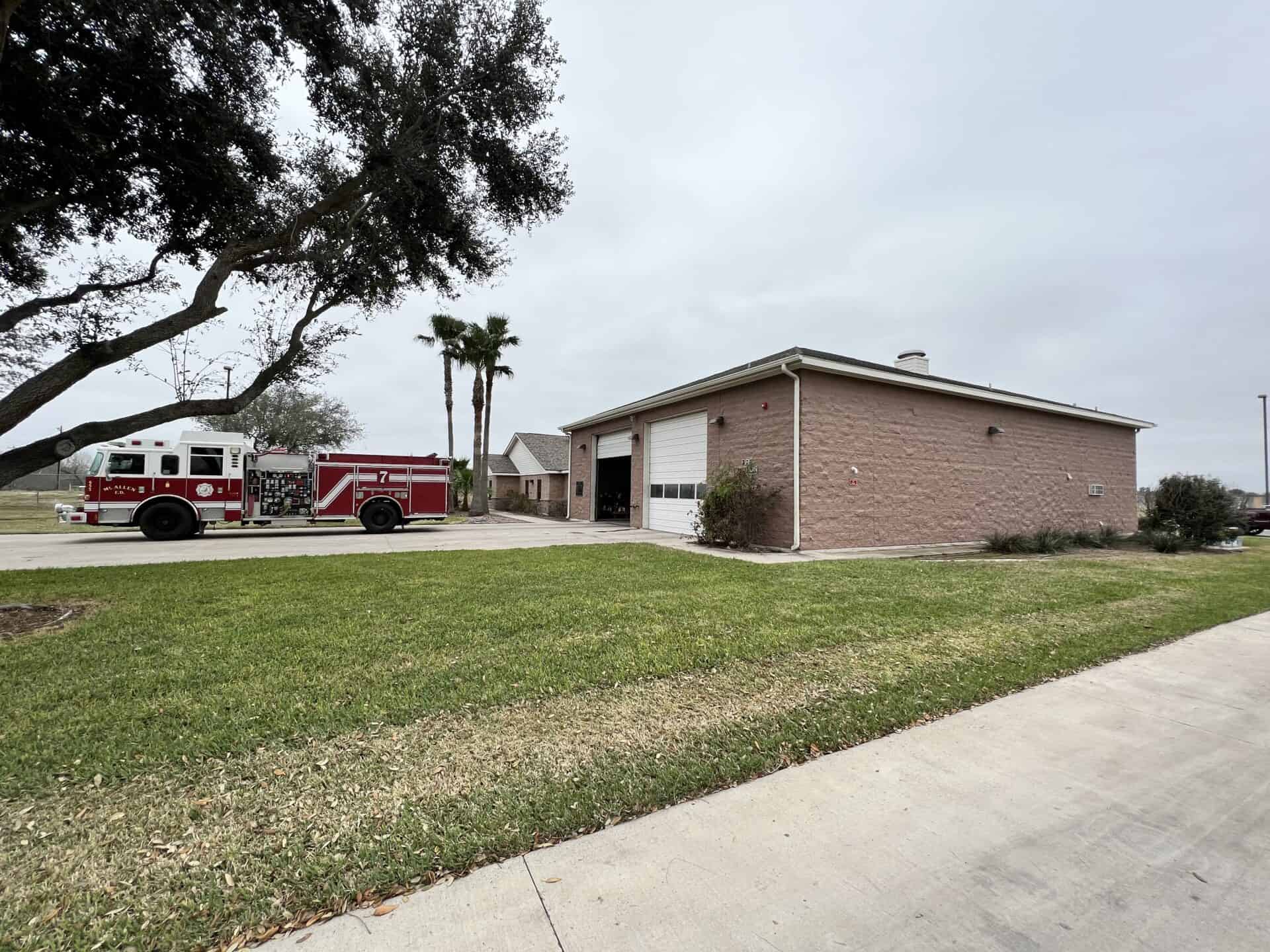 A red fire truck is parked outside a beige brick fire station on a cloudy day. The area is surrounded by green grass, trees, and a smooth driveway.