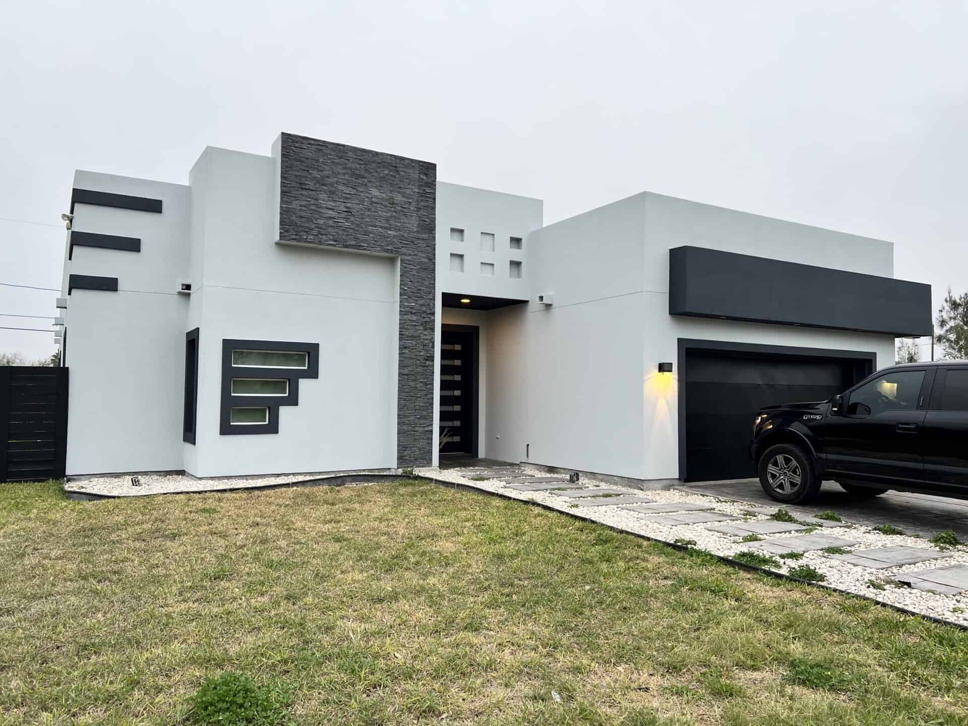 Modern white house with geometric architecture, gray stone accents, and horizontal windows. A black truck is parked on a stone driveway. Overcast sky.