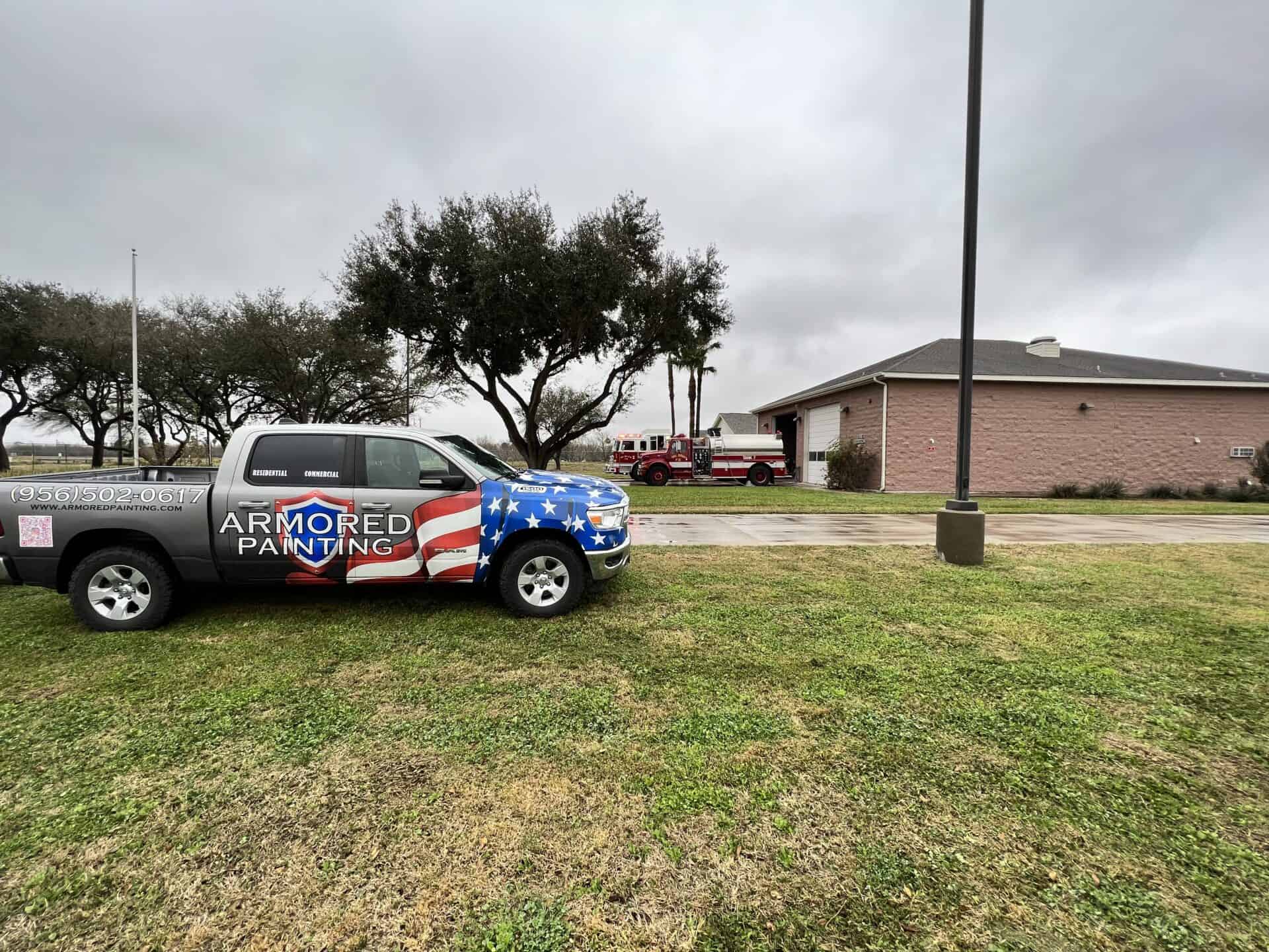 Gray sky over a grassy field with a truck featuring an American flag design and logo reading "Armored Painting." Nearby, a pink building and trees add contrast.