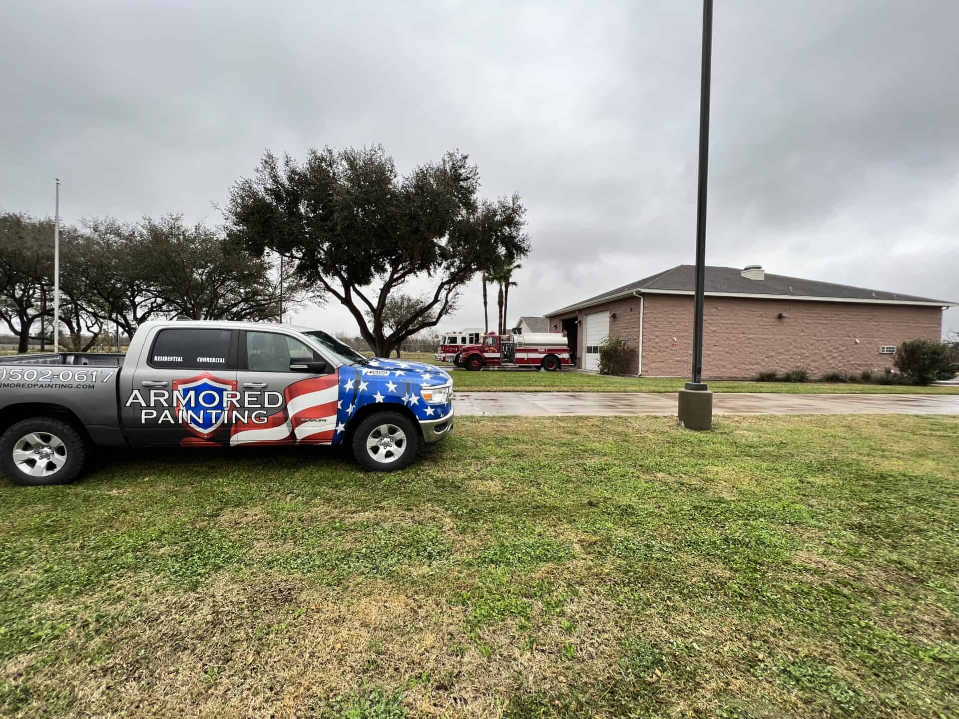 A pickup truck with an American flag design labeled "Armored Painting" is parked on grass near a pink building and a red firetruck under a cloudy sky.