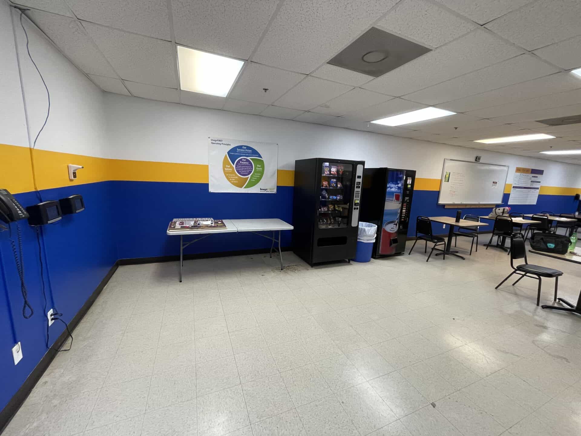 Break room with blue and yellow walls, vending machines, and empty tables. Fluorescent lights create a quiet, utilitarian atmosphere.