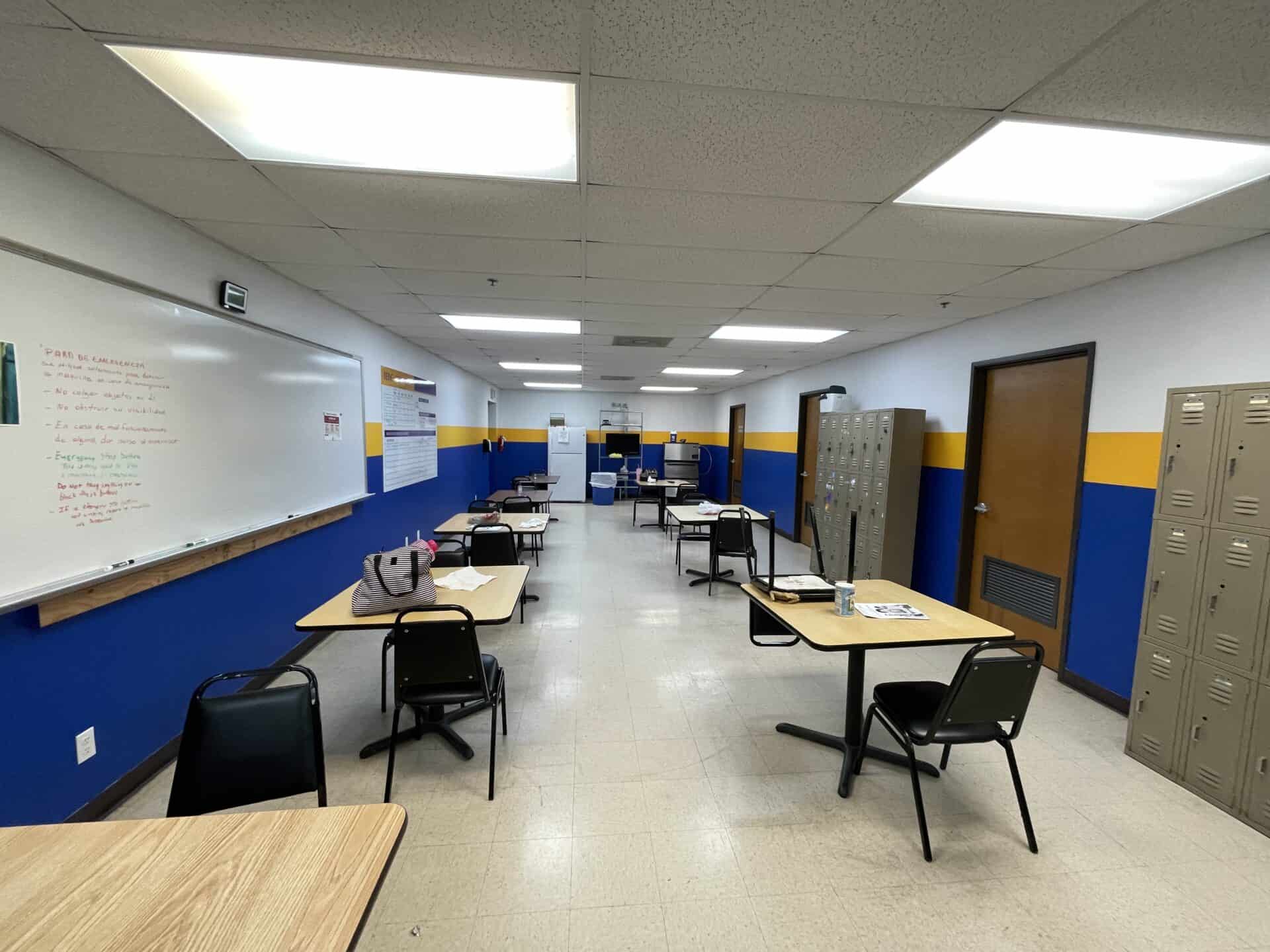 Classroom with blue and yellow walls features tables and chairs in a row, a whiteboard with notes, and lockers lining one side. Bright, organized, and tidy.