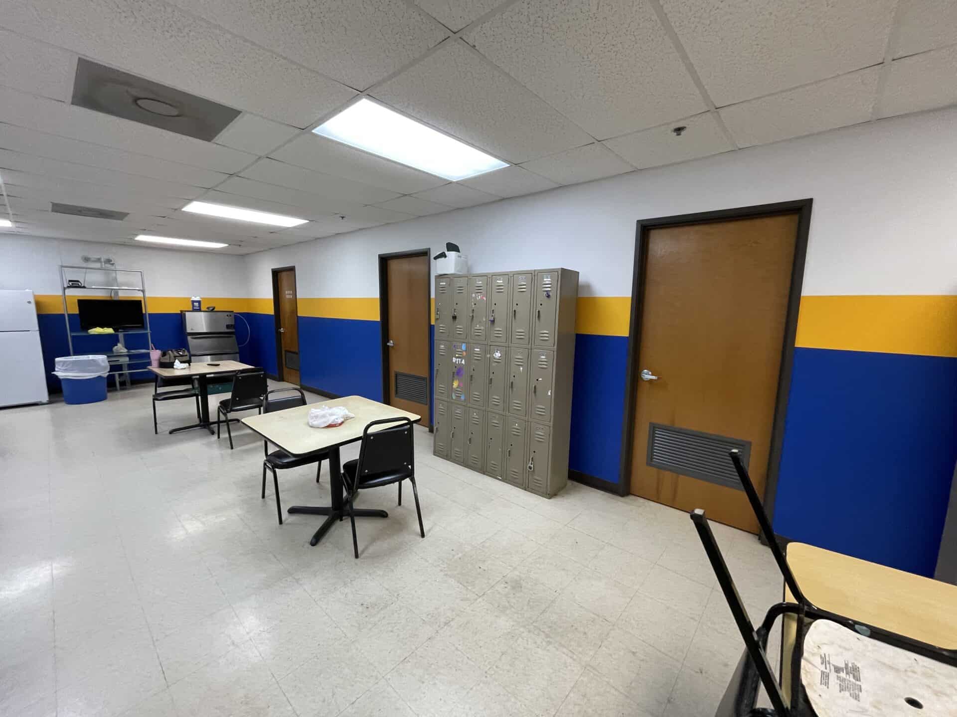 Empty break room with yellow and blue walls, two tables with chairs, lockers, fridge, and shelves. Fluorescent lights create a functional ambiance.
