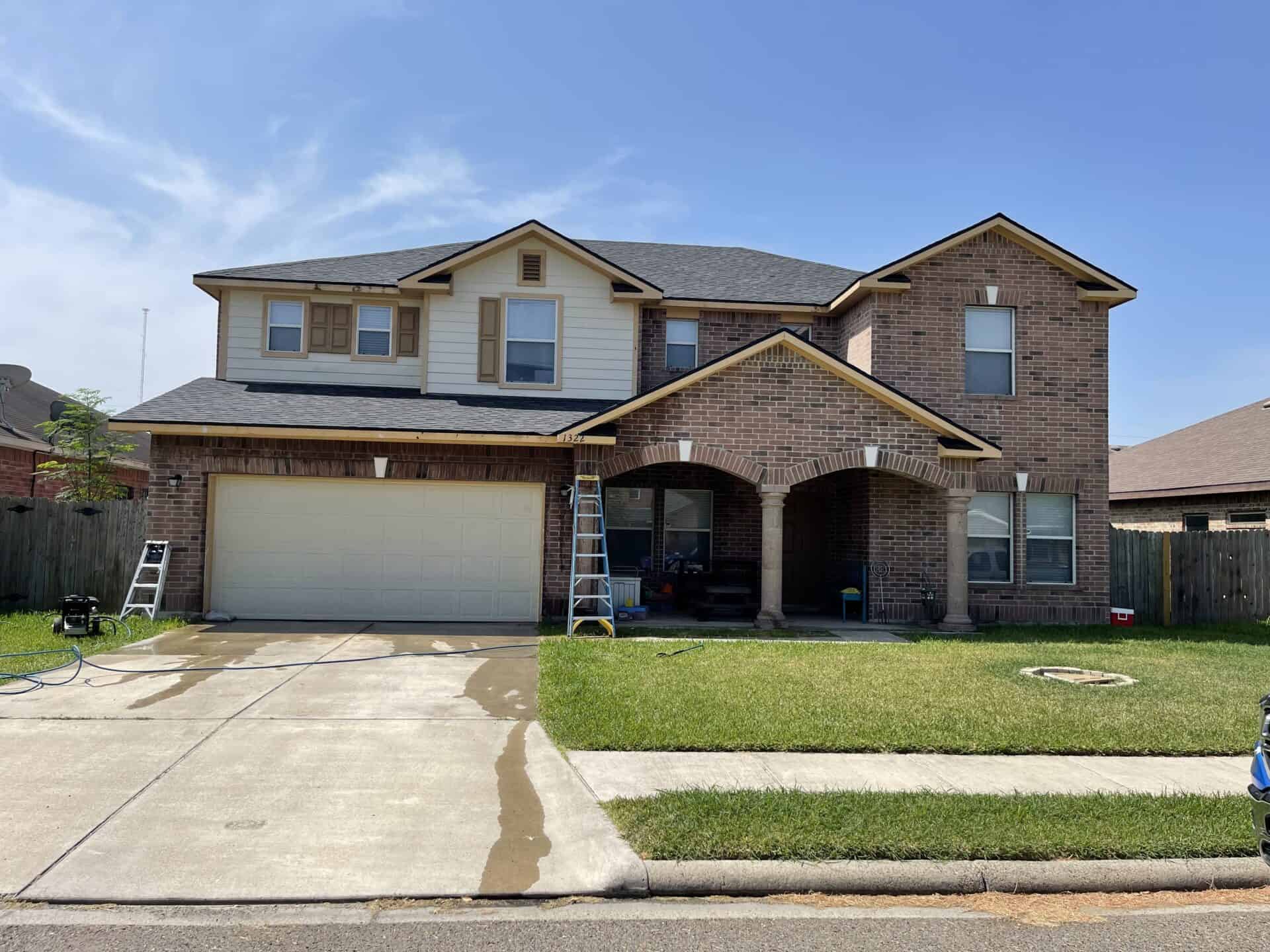 Two-story brick house with a two-car garage and an arched front porch. Two ladders are set up outside, and the driveway shows signs of cleaning.