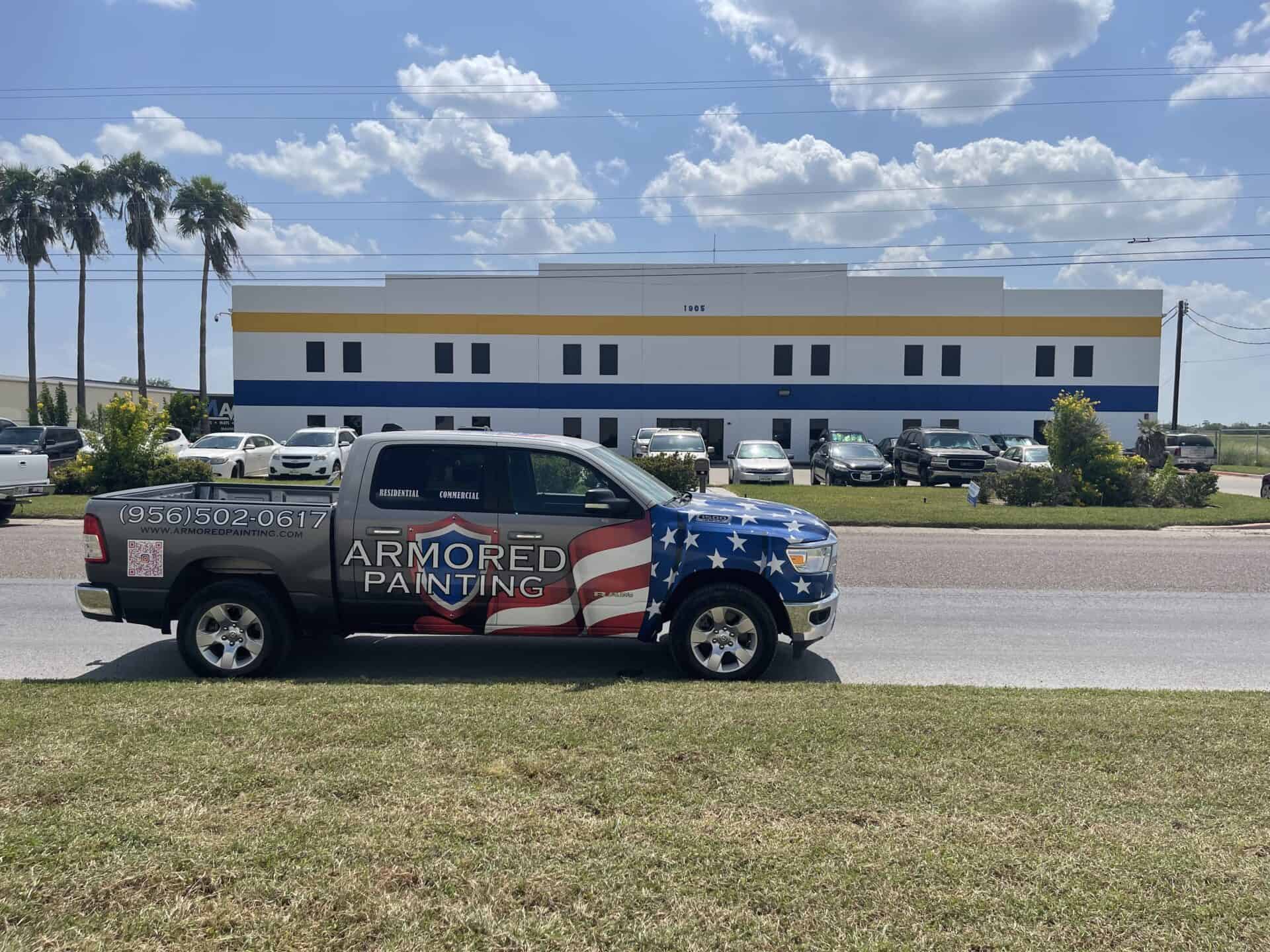 A truck with a patriotic design and "Armored Painting" logo is parked on a road. A modern building and palm trees are in the background under a bright blue sky.