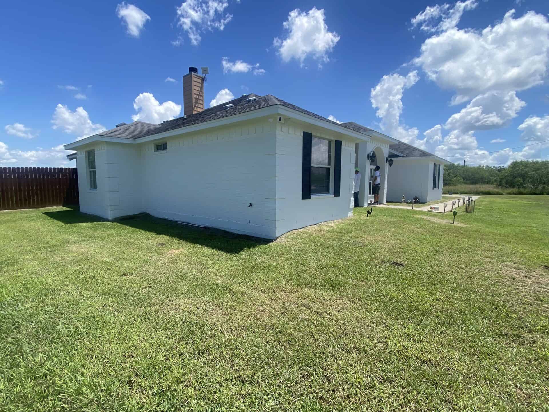Single-story white house with black shutters, surrounded by an expansive green lawn under a bright blue sky with fluffy clouds, creating a serene atmosphere.