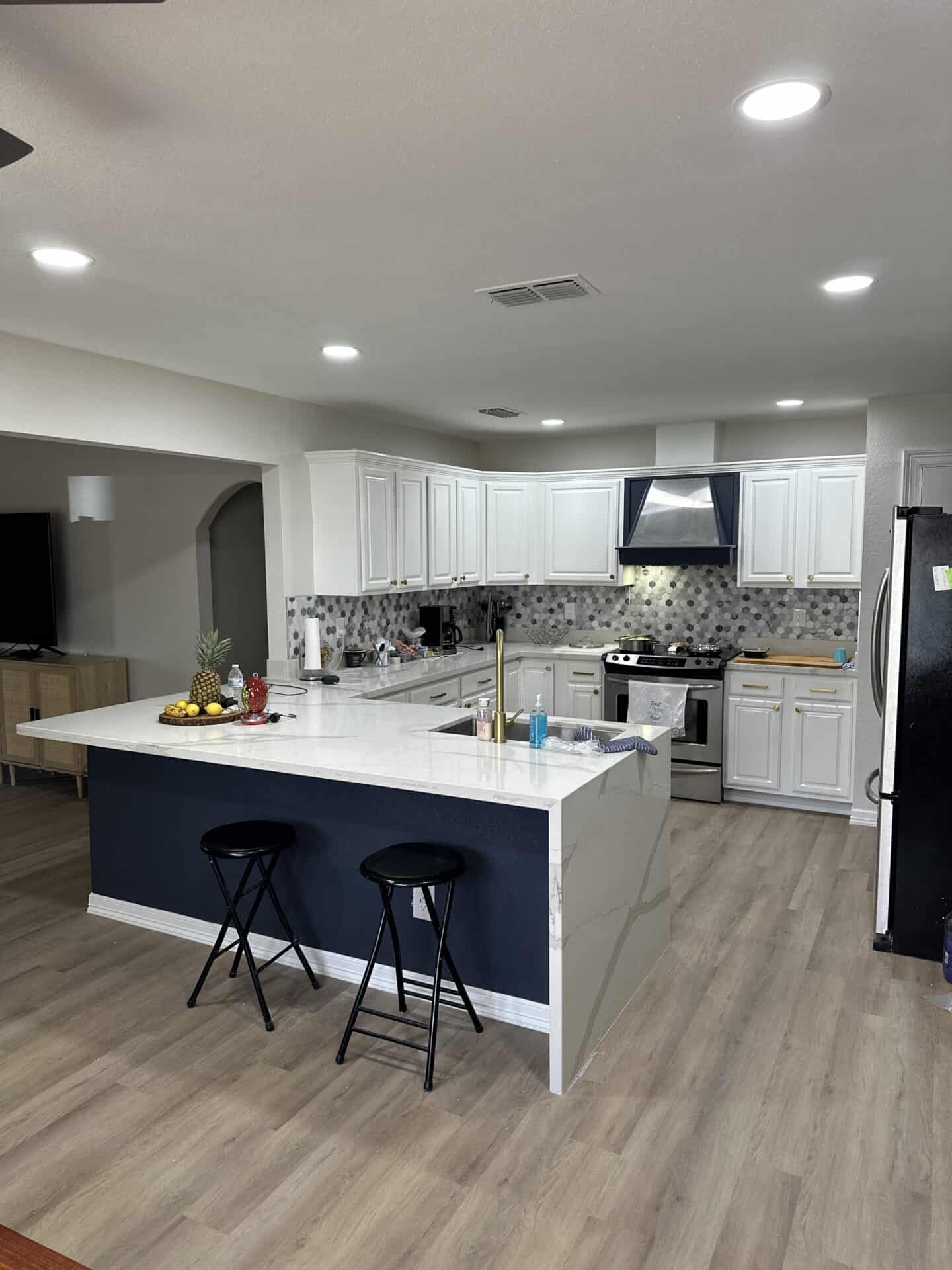 Modern kitchen with white cabinets, marble countertops, and stainless steel appliances. An island with two black stools and fruit bowl adds warmth.