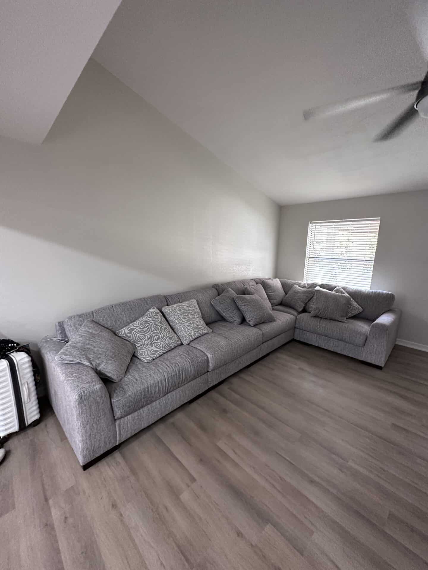 Spacious living room with a gray L-shaped couch, patterned throw pillows, wooden floor, white walls, and a window with blinds. Calm and inviting ambiance.
