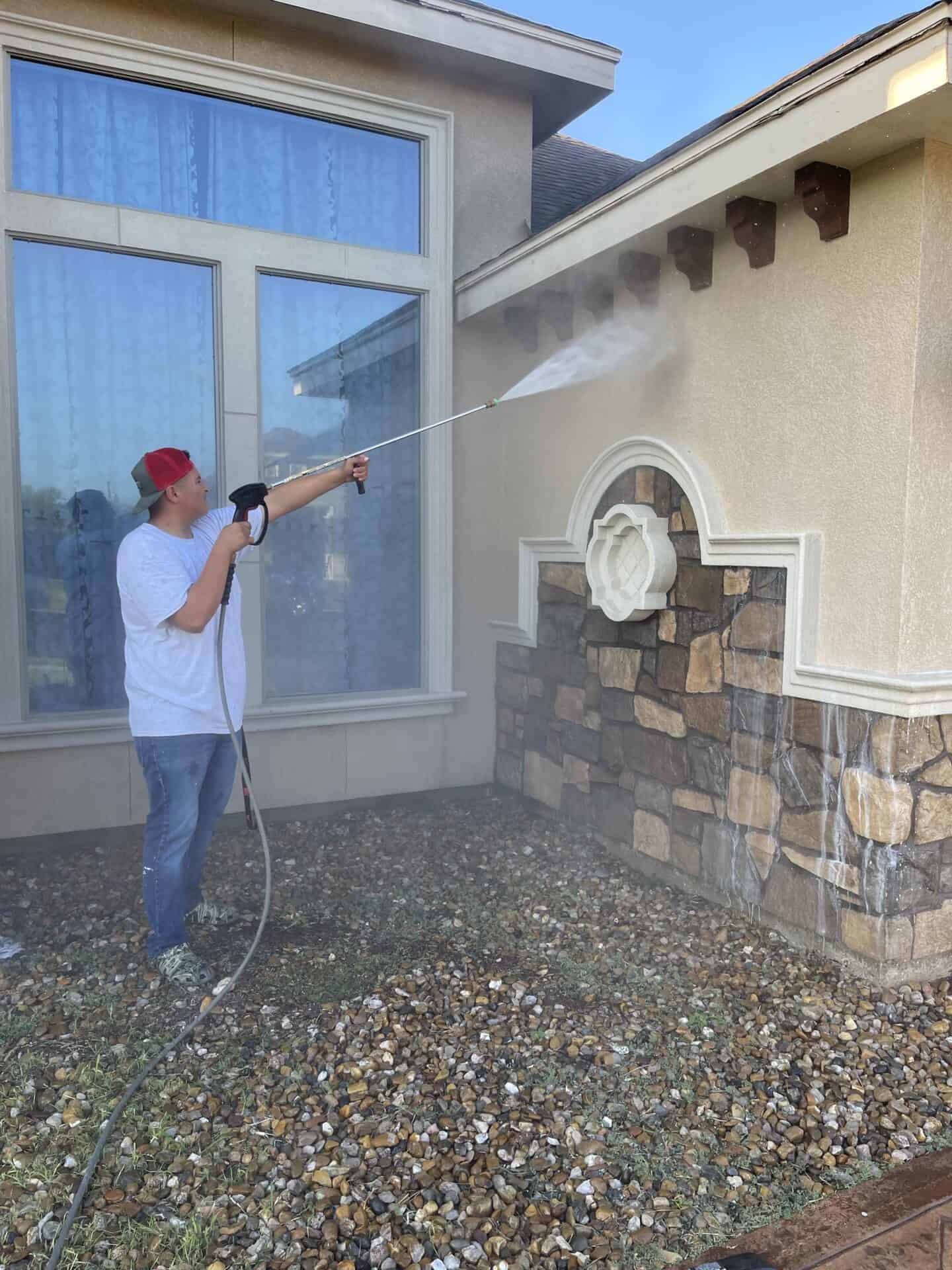 Man in red cap pressure washing a stone wall next to large windows. Water sprays forcefully on the wall, cleaning dirt. Bright, clear day.