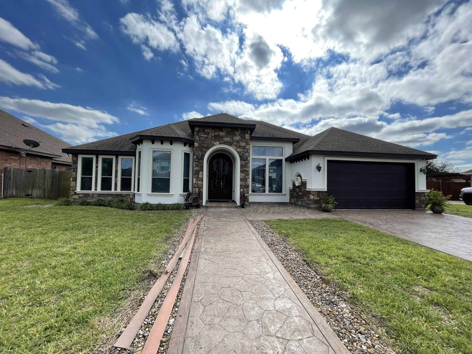 Modern, single-story house with a stone archway entrance, large windows, and a dark garage. The day is partly cloudy with a vibrant blue sky.