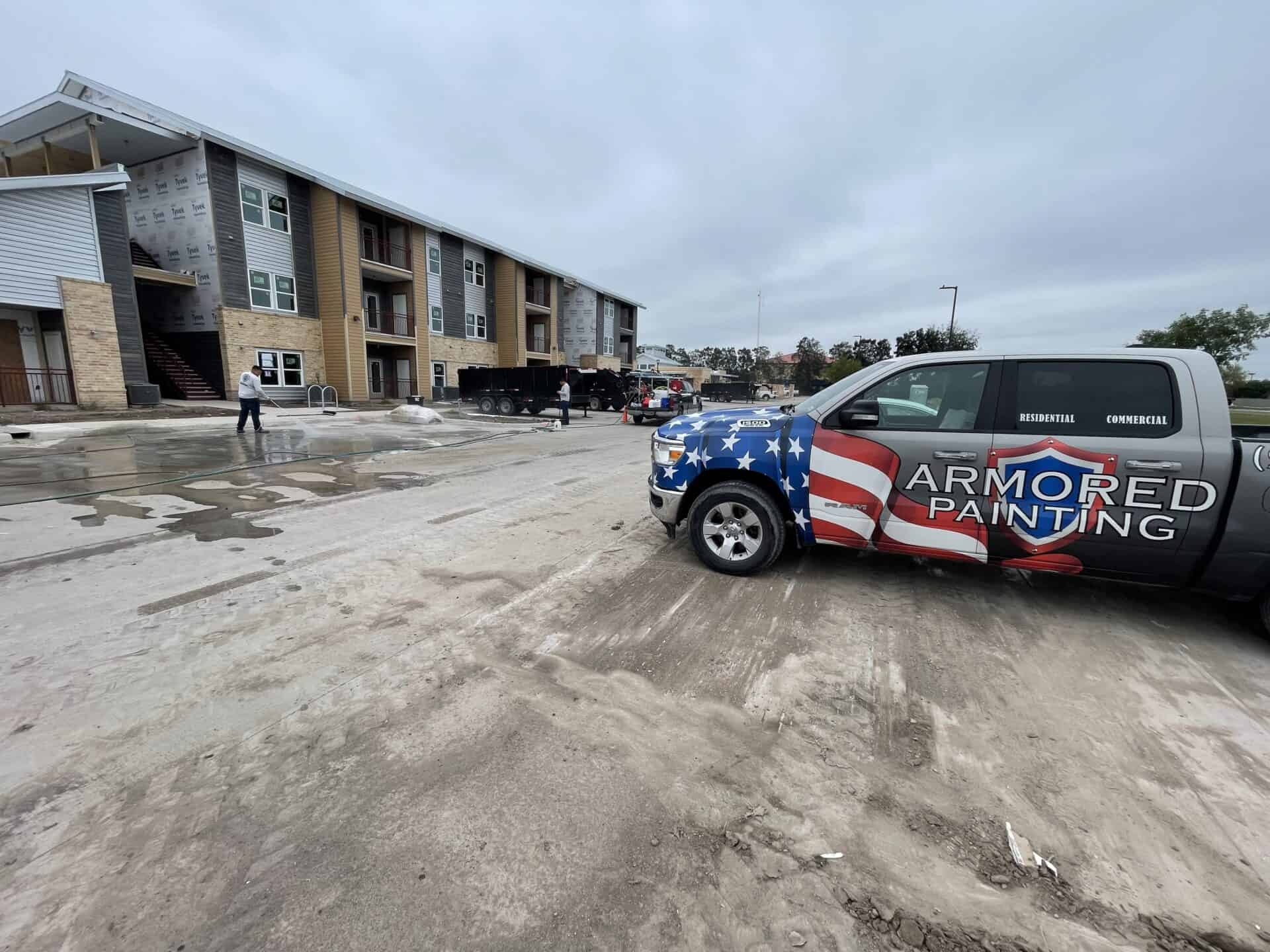 Armored Painting truck with American flag design parked at construction site in front of a partially built apartment complex under gray sky. Workers nearby.
