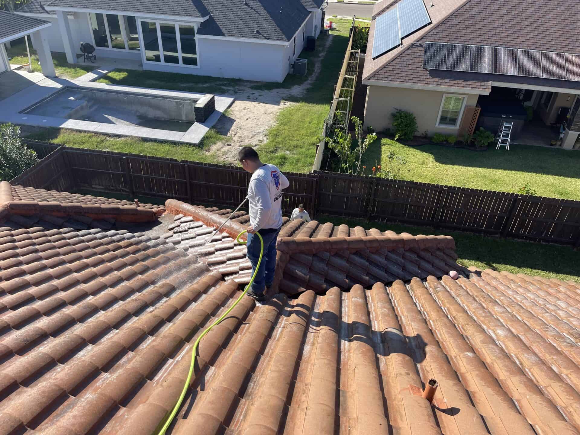 Man in blue jeans and a white shirt pressure washes a terracotta-tiled roof. Surrounding homes have solar panels and a pool, under a clear sky.