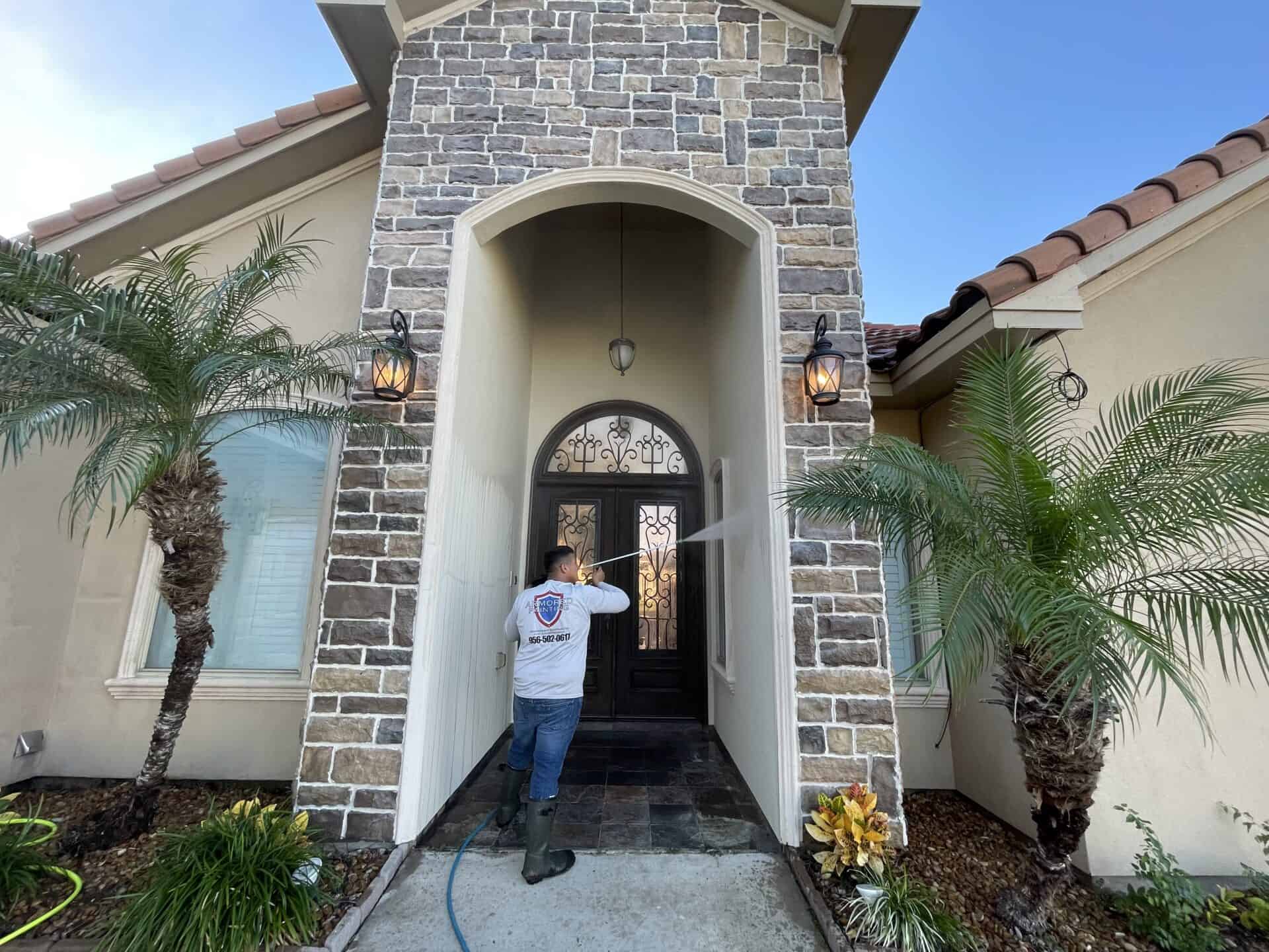 Worker power-washing stone exterior of a house with palm trees flanking the entrance. The scene conveys outdoor maintenance and cleanliness.